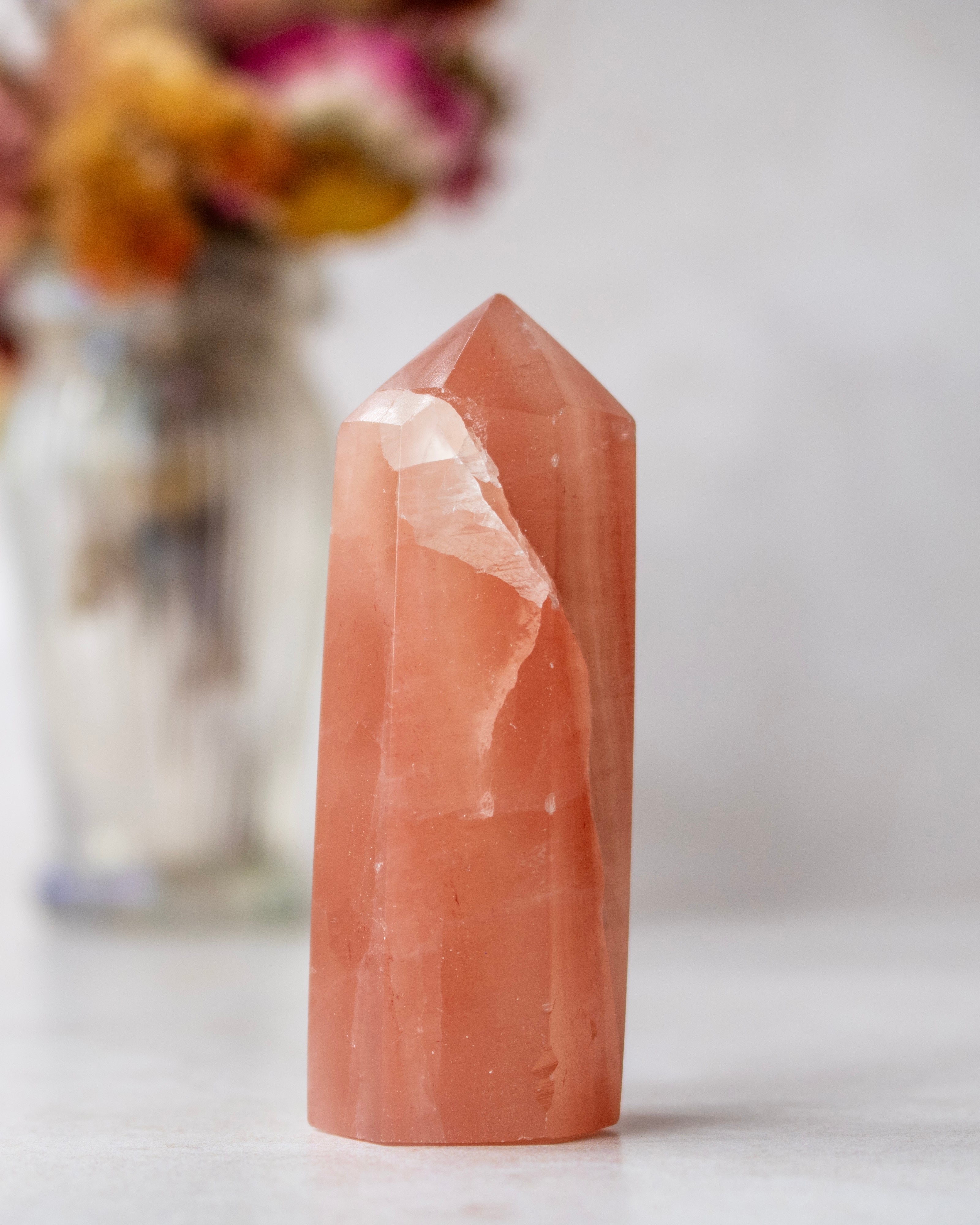 A natural peach-colored quartz crystal point tower standing upright on a light surface, with a softly blurred glass vase of dried flowers in the background.