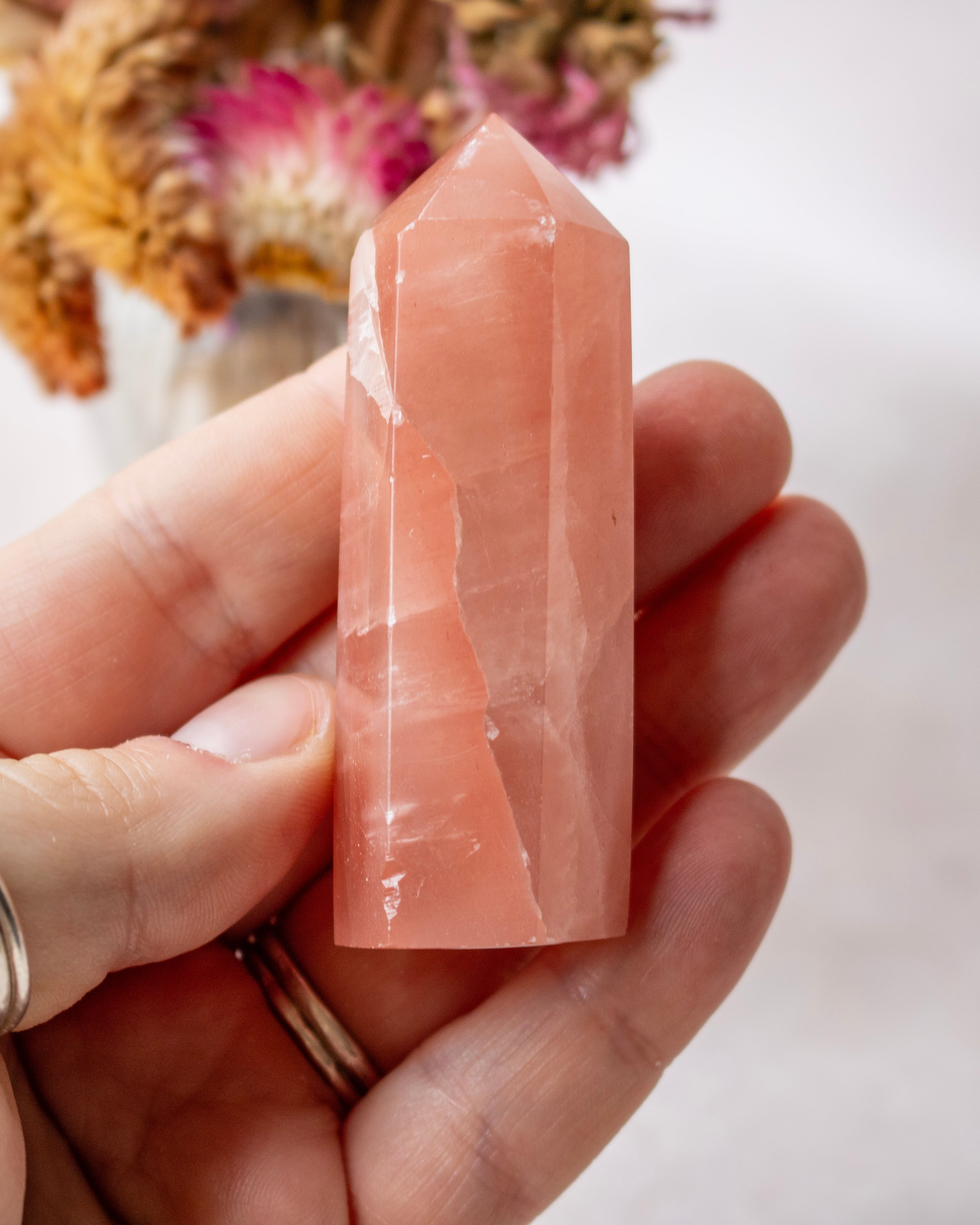 A hand holding a polished rose quartz crystal point against a soft, blurred background with dried flowers.
