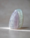 A polished iridescent crystal stone with soft lavender, pale blue, and mint green tones, standing upright on a neutral gray surface with a soft shadow.