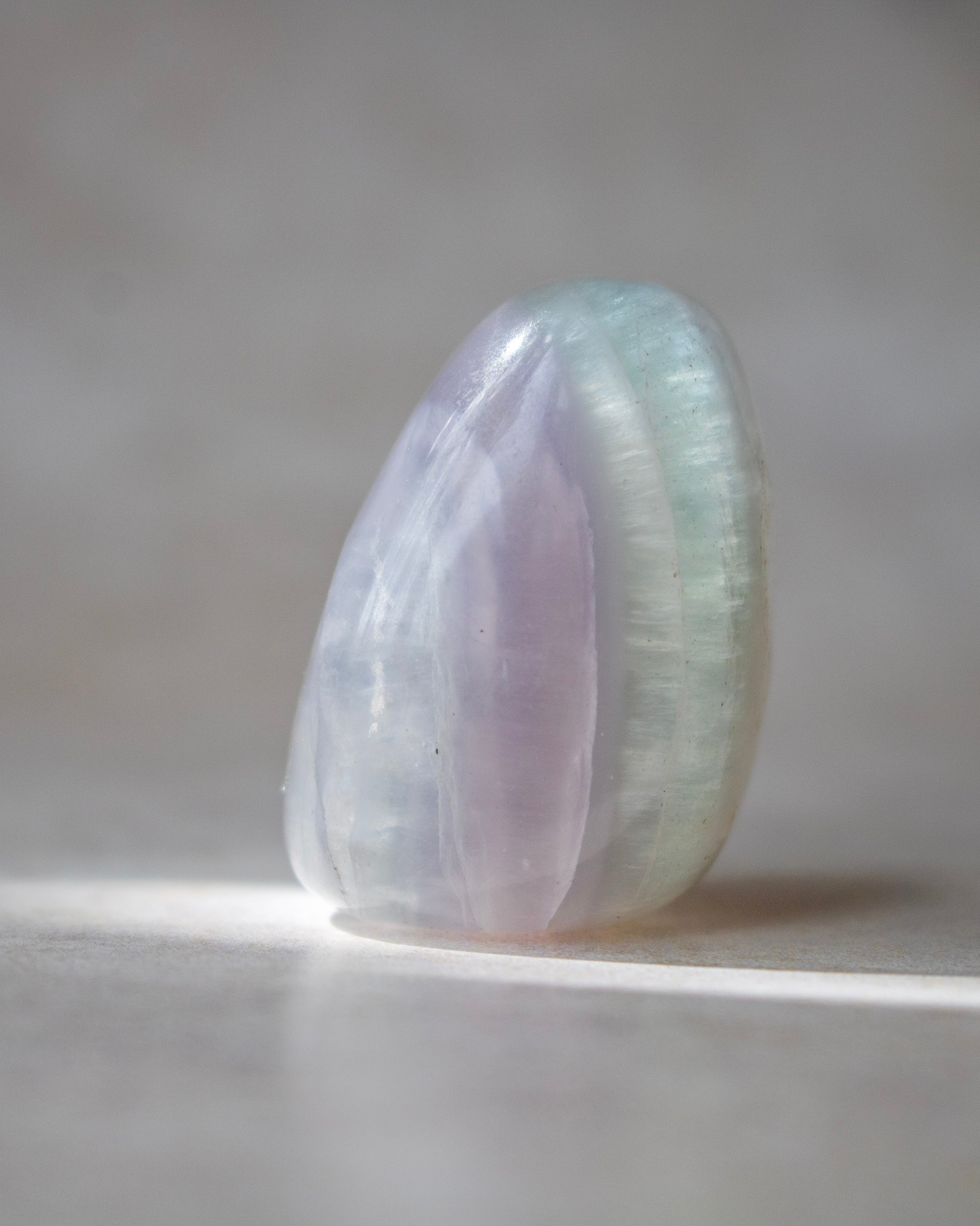 A polished iridescent crystal stone with soft lavender, pale blue, and mint green tones, standing upright on a neutral gray surface with a soft shadow.