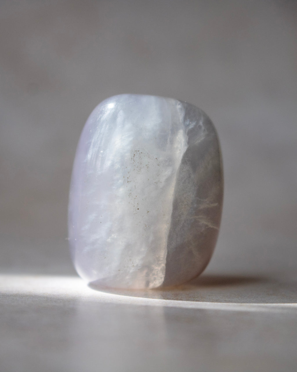 A polished tumbled crystal stone with a rounded rectangular shape, featuring a milky white body with subtle lavender-gray veining and a smooth, reflective surface, standing on a neutral surface against a soft gray background.