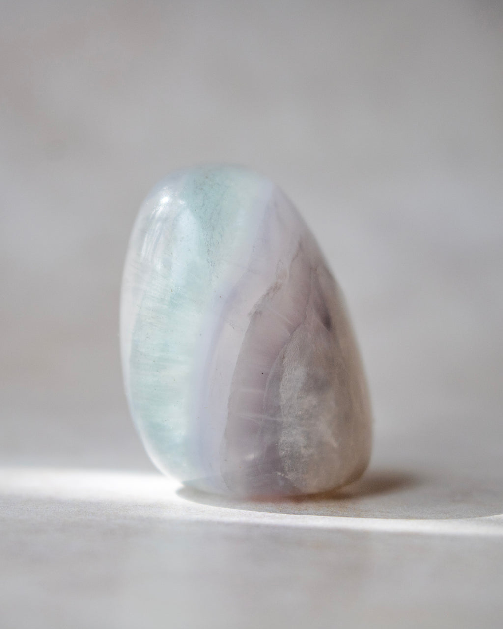 A polished tumbled crystal stone with soft pastel bands of pale blue-green, lavender, and gray, standing on a light neutral surface.
