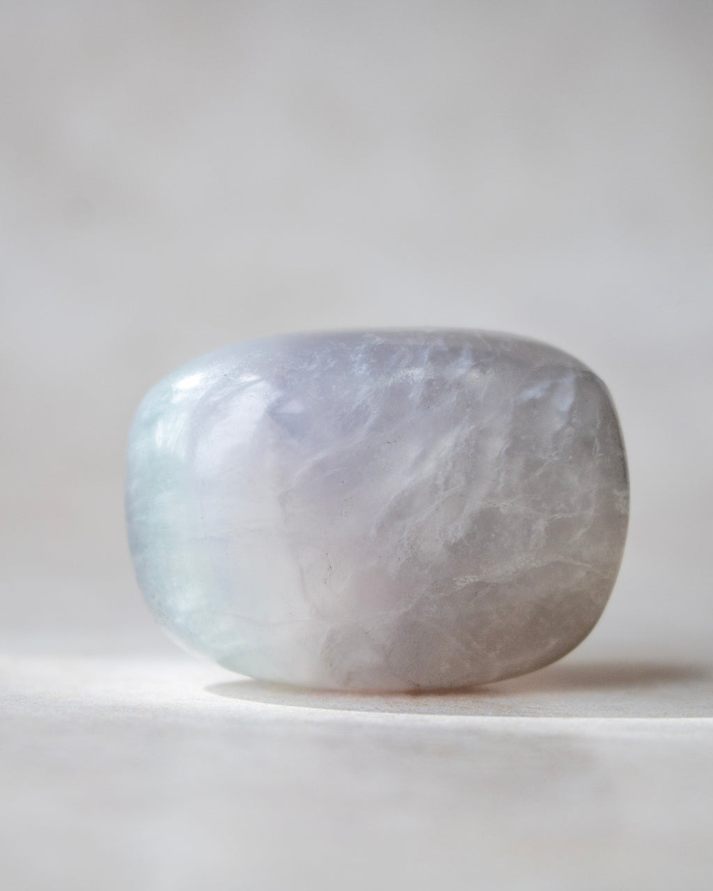 A polished, rounded crystal stone with a smooth, matte finish, displaying a soft lavender-gray color with subtle white and pale blue marbling, resting on a neutral light gray surface.