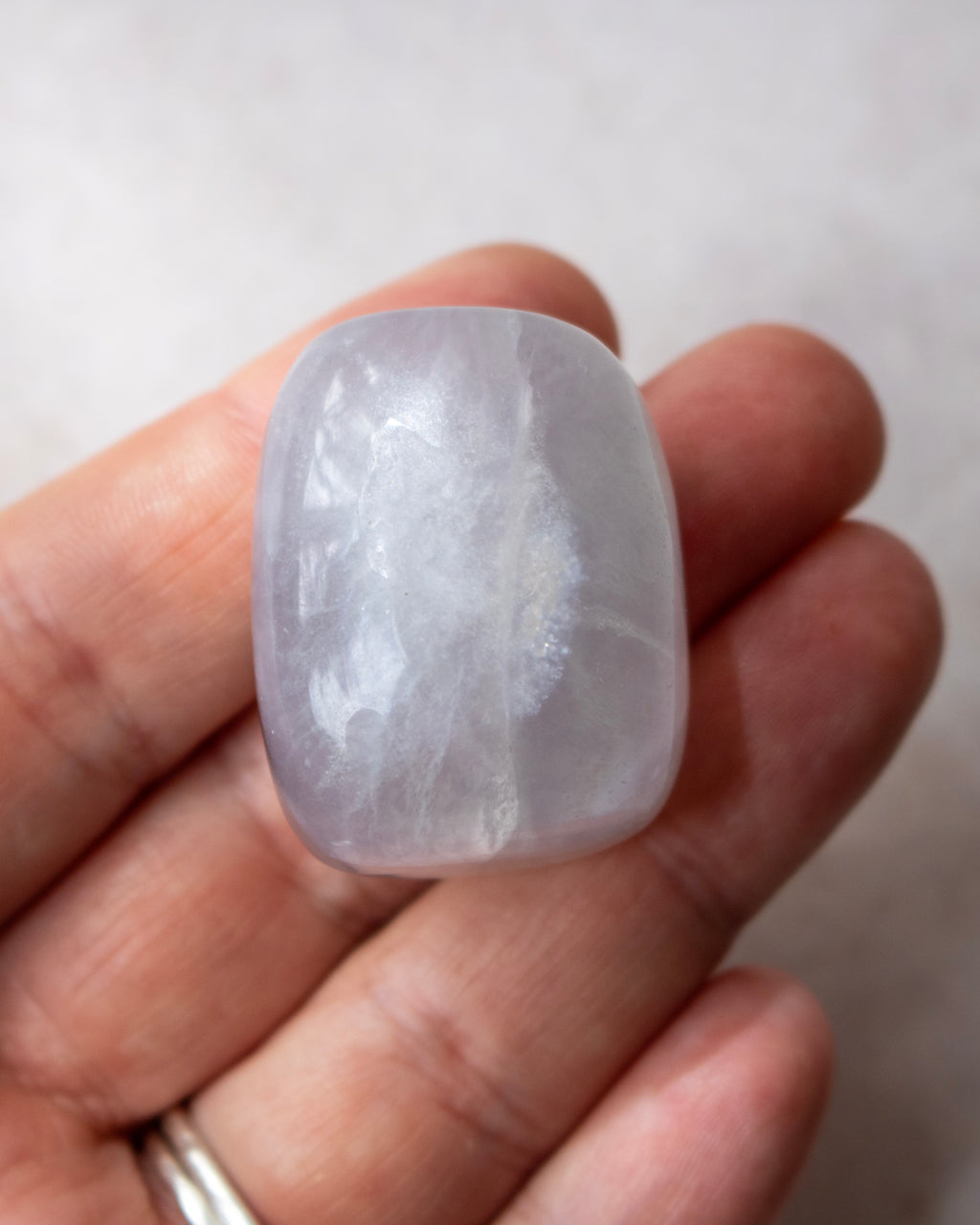 A hand holding a smooth, polished white stone cabochon with subtle grayish-purple undertones and natural inclusions, set against a soft-focus light gray background.