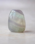 A polished tumbled stone with iridescent bands of pale green, lavender purple, and soft gray, standing upright on a light beige surface against a soft neutral background.