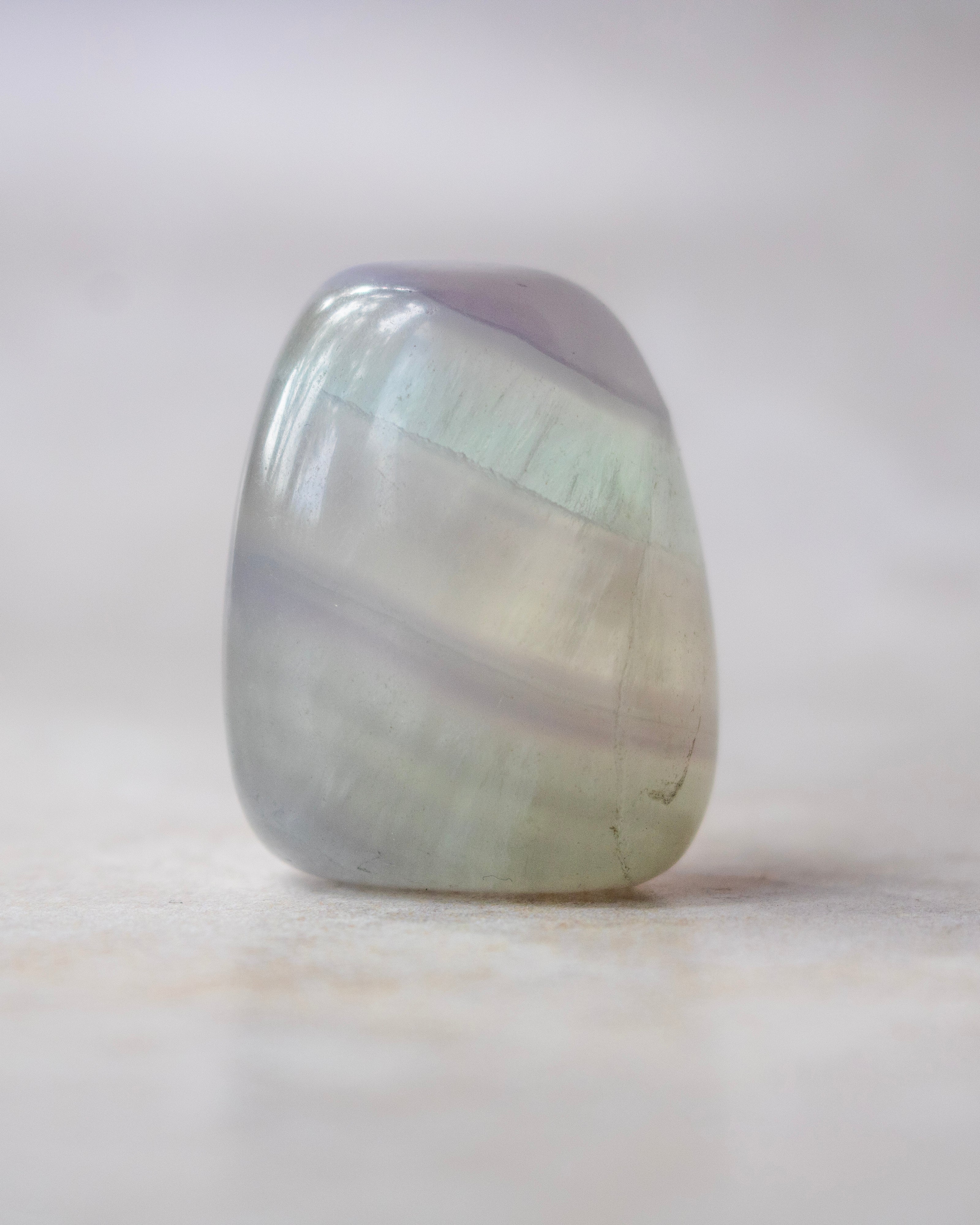 A polished tumbled stone with iridescent bands of pale green, lavender purple, and soft gray, standing upright on a light beige surface against a soft neutral background.