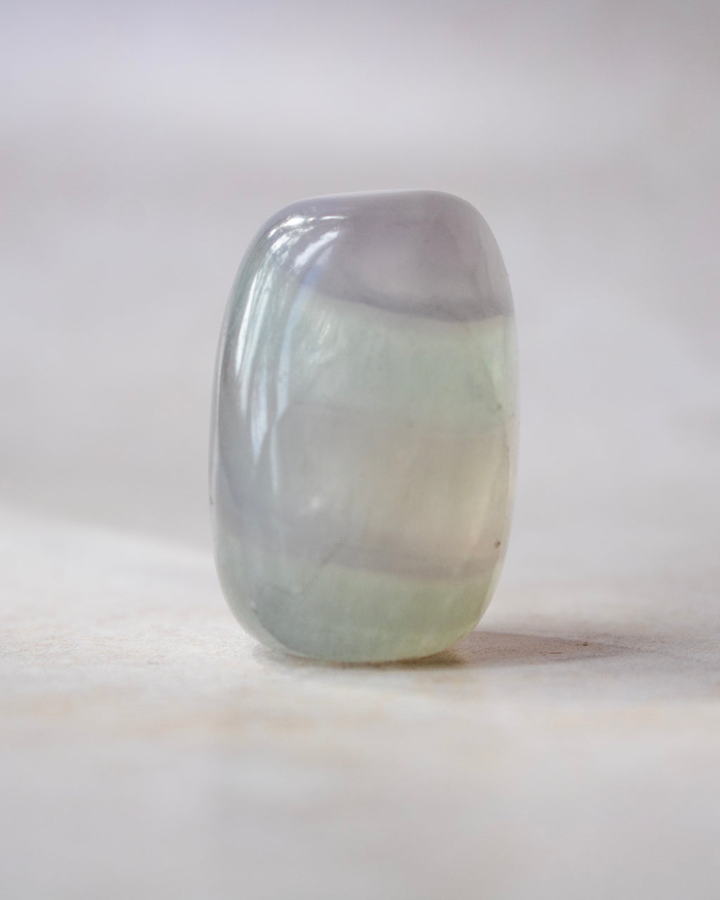 A polished, rounded rectangular gemstone with a smooth, glossy finish, featuring a color gradient from pale lavender-gray at the top to a soft minty green at the bottom, standing upright on a light beige surface.