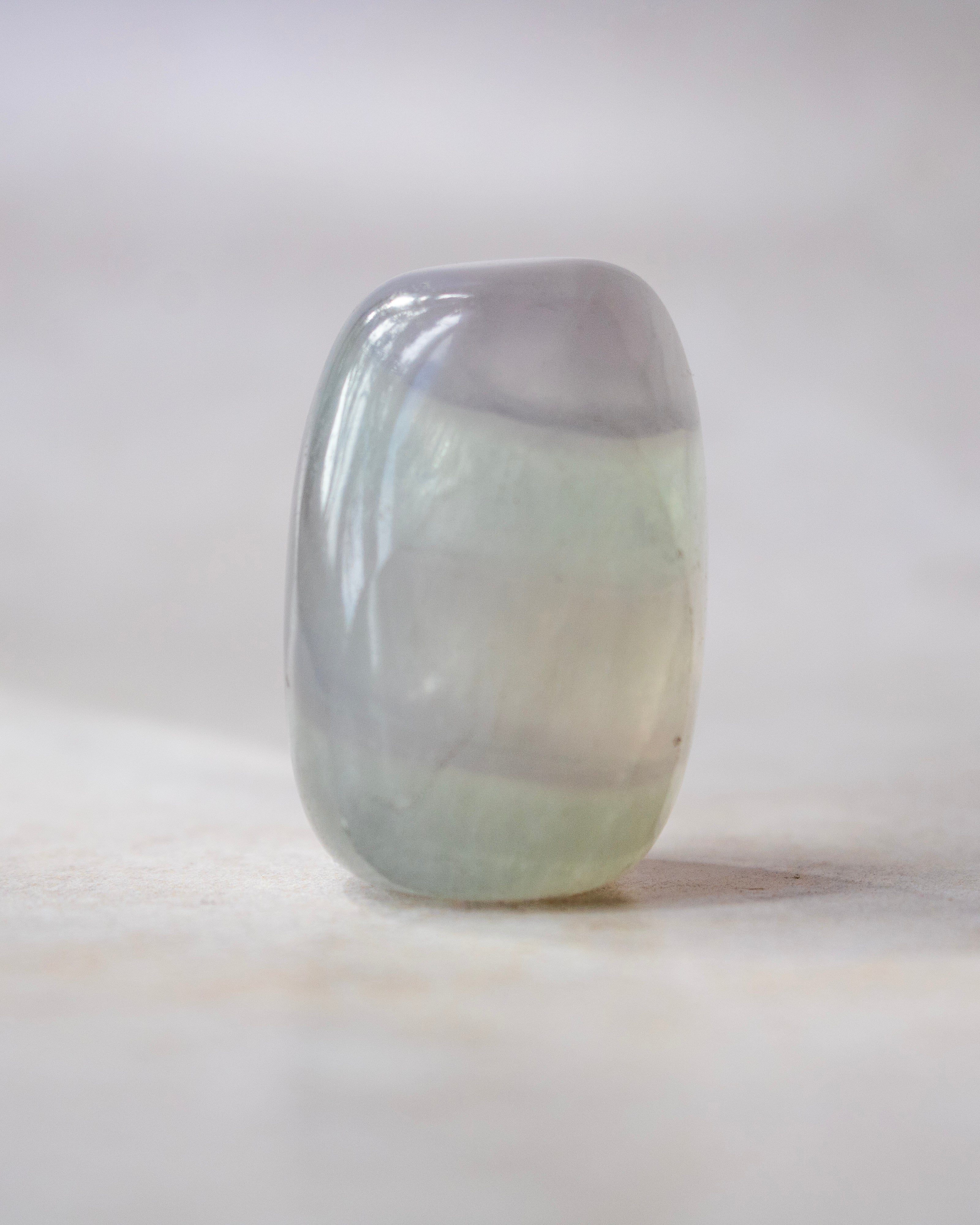 A polished, rounded rectangular gemstone with a smooth, glossy finish, featuring a color gradient from pale lavender-gray at the top to a soft minty green at the bottom, standing upright on a light beige surface.