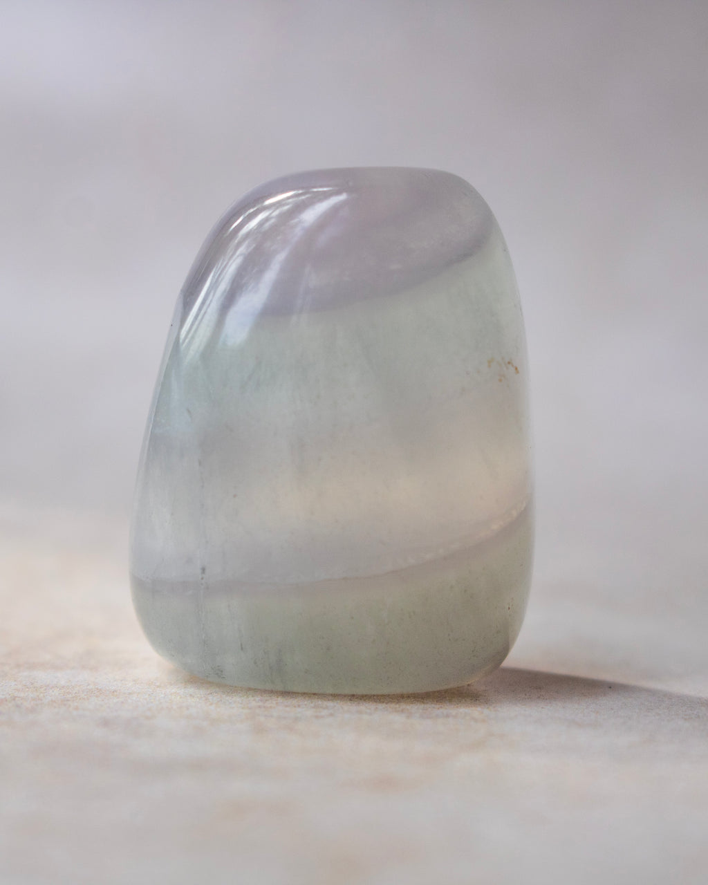A polished, teardrop-shaped crystal with a smooth, glossy finish, showcasing a color transition from pale lavender-gray at the top to light green at the bottom, resting on a neutral beige surface.