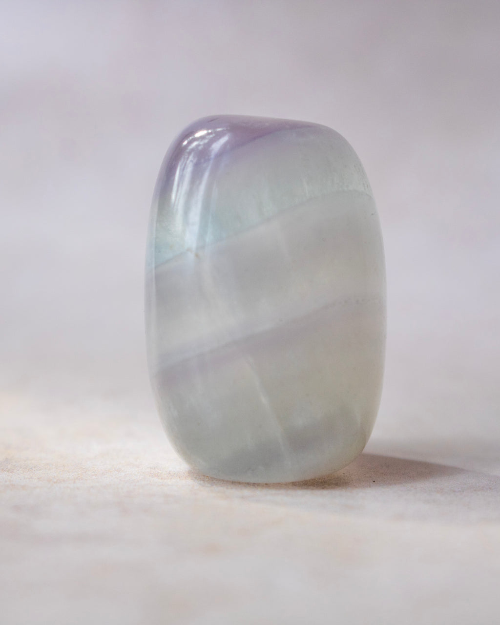 A polished gemstone tumbled into a rounded bar shape, featuring iridescent bands of lavender, pale green, and white, set against a soft, light beige background.