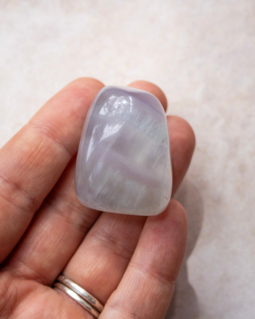 A hand holding a smooth, polished light purple tumbled stone, against a neutral light beige background.
