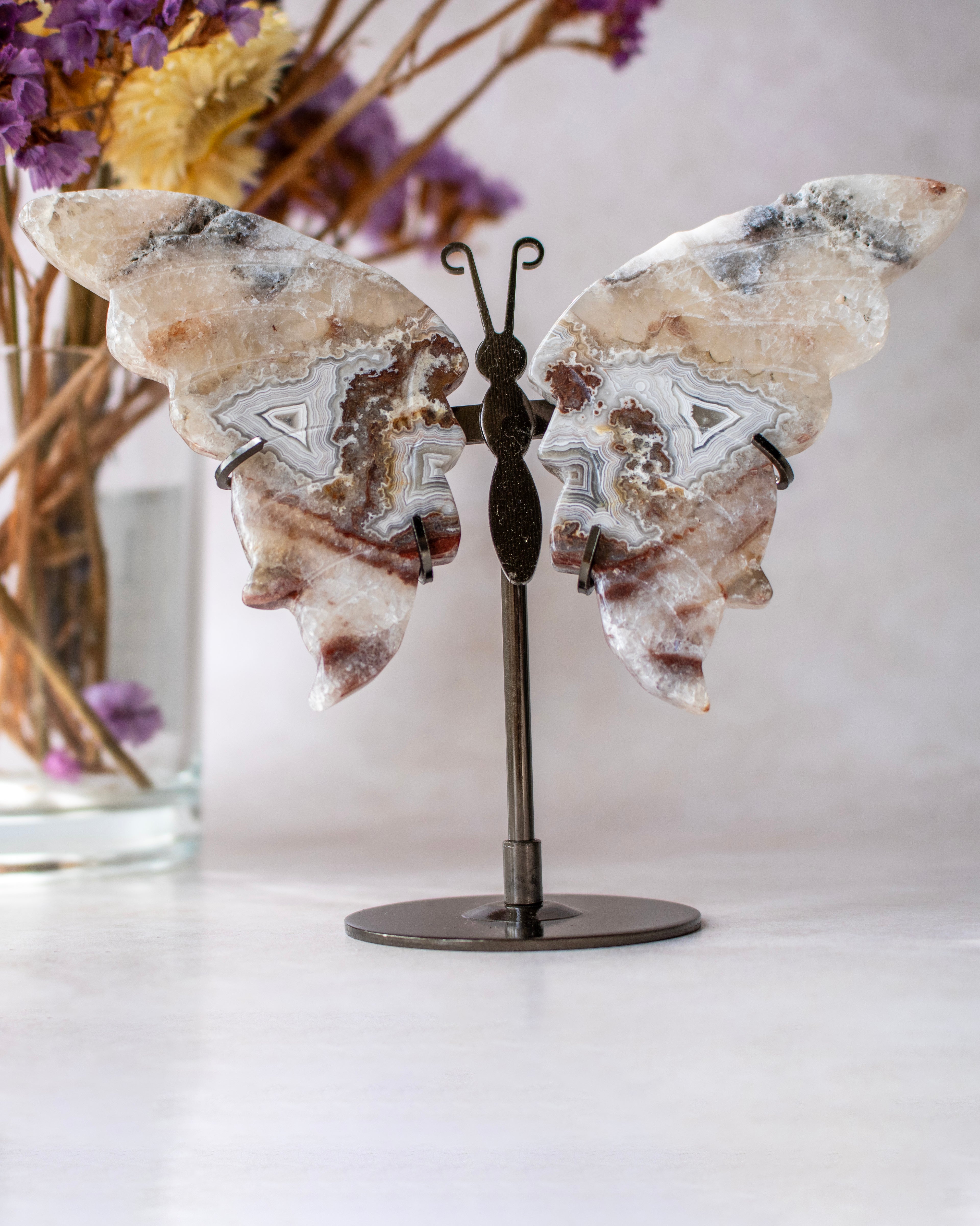 A butterfly-shaped crystal cluster display stand with two natural agate wings and a dark metal stand, shown on a light surface with dried flowers in the background.