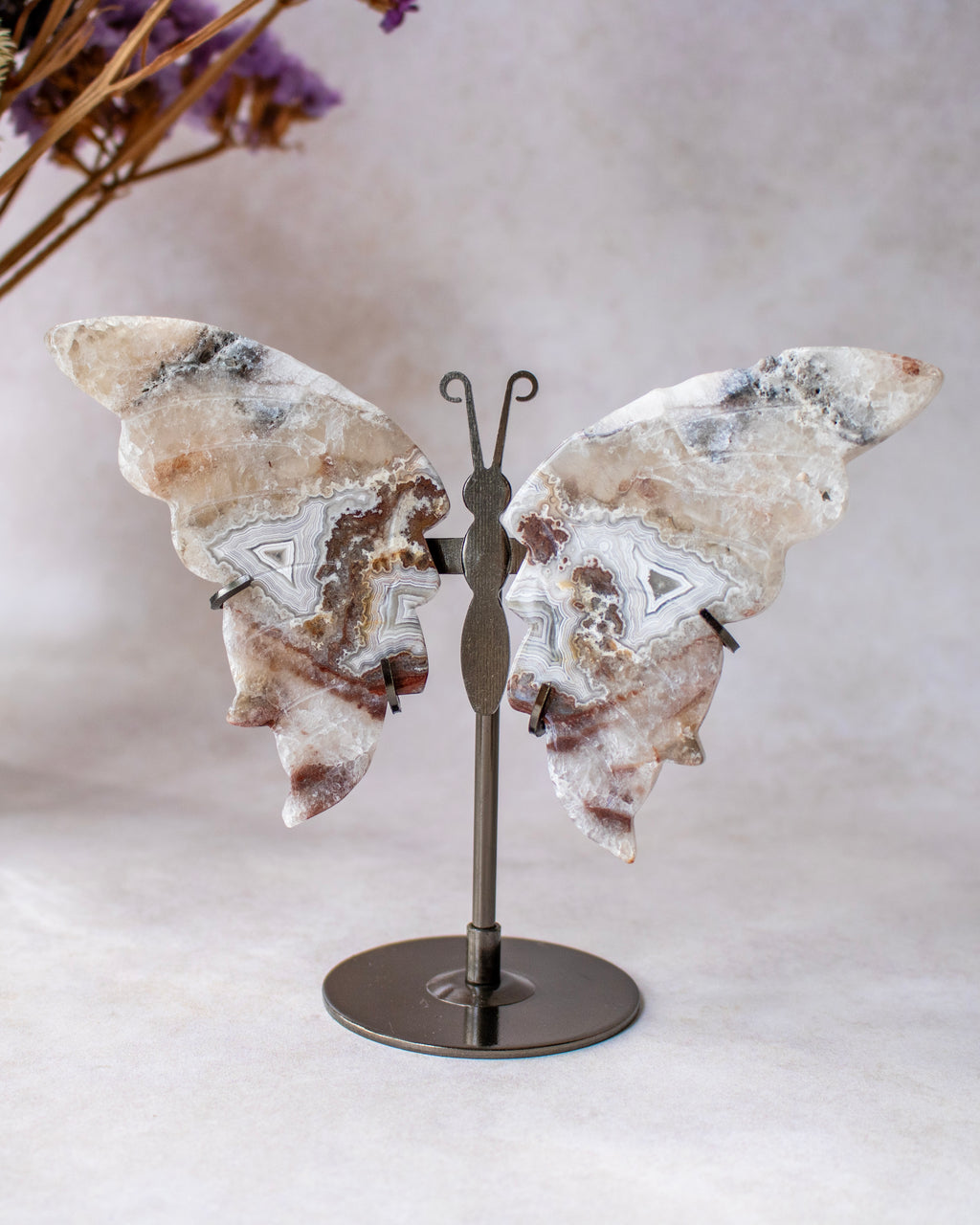 A butterfly-shaped crystal sculpture made from agate with natural brown, gray, and white veining, mounted on a dark metal stand against a neutral light gray background.