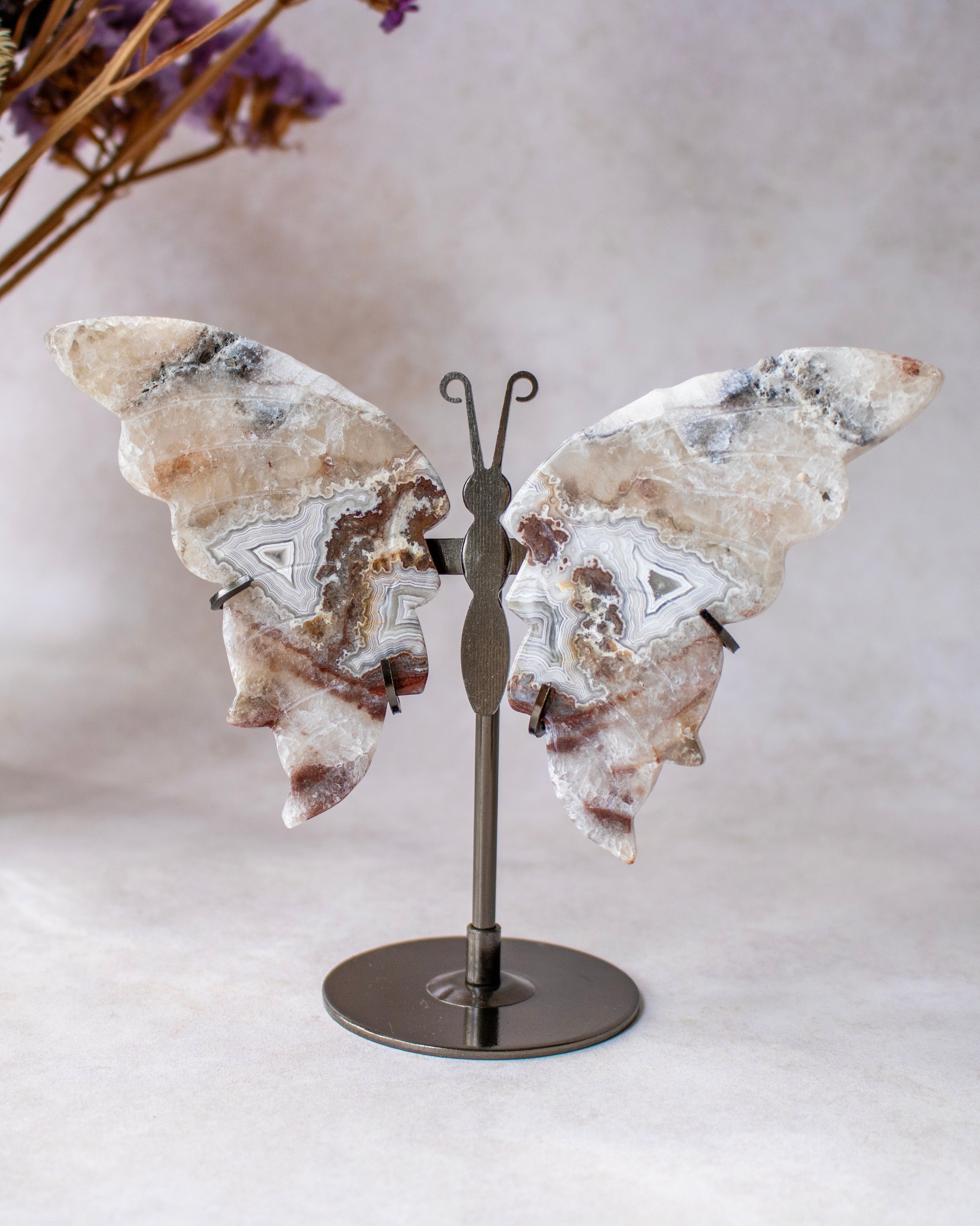 A butterfly-shaped crystal sculpture made from agate with natural brown, gray, and white veining, mounted on a dark metal stand against a neutral light gray background.