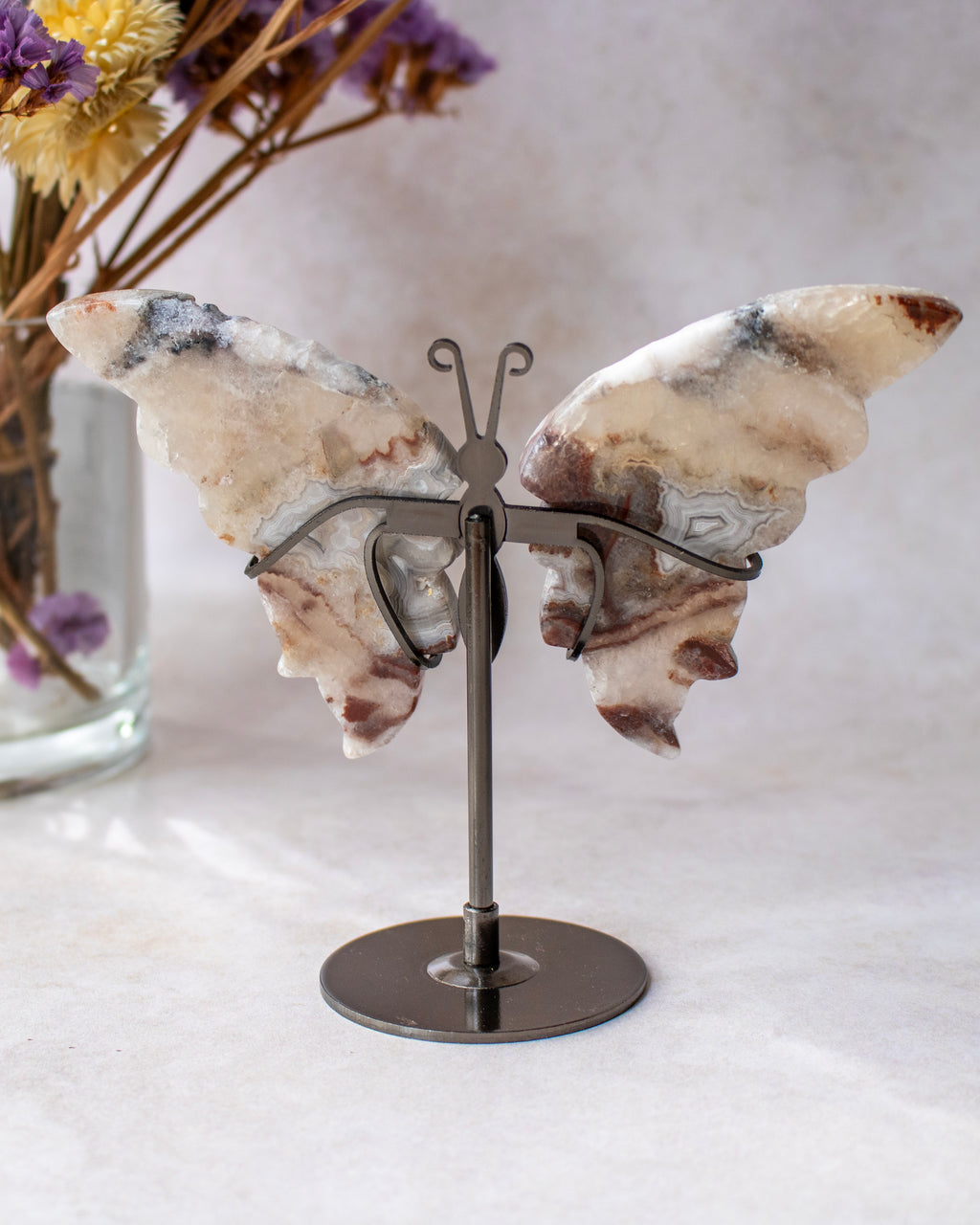 A butterfly-shaped wall art piece made from natural agate wings in cream, beige, and brown tones, mounted on a dark metal stand with a circular base, set against a soft neutral background with dried flowers in the side.