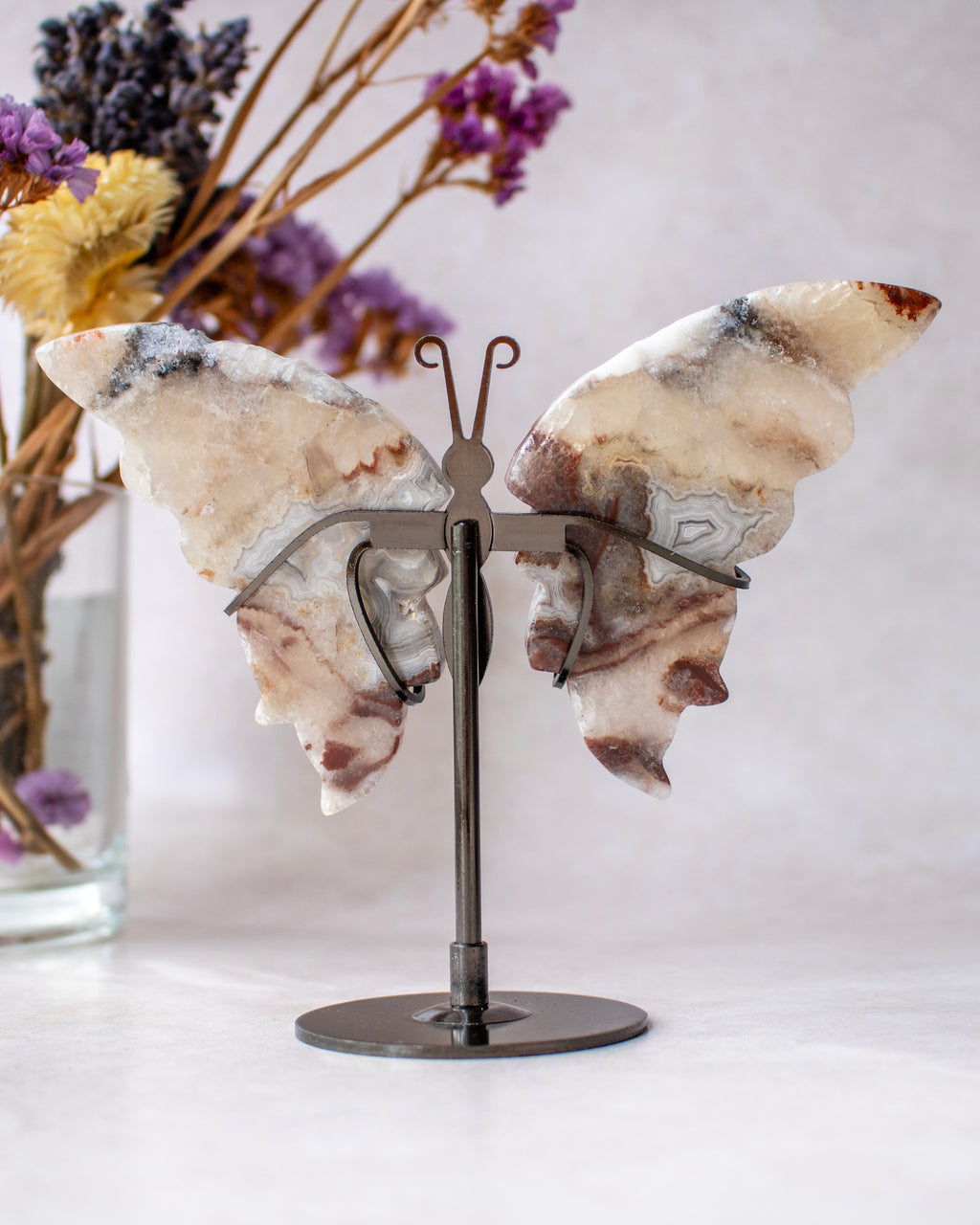 A butterfly-shaped wall art piece made from natural agate with marbled beige, white, and brown patterns, mounted on a dark metal stand. The stand is placed on a light surface with a soft-focus background showing dried flowers in a glass vase.