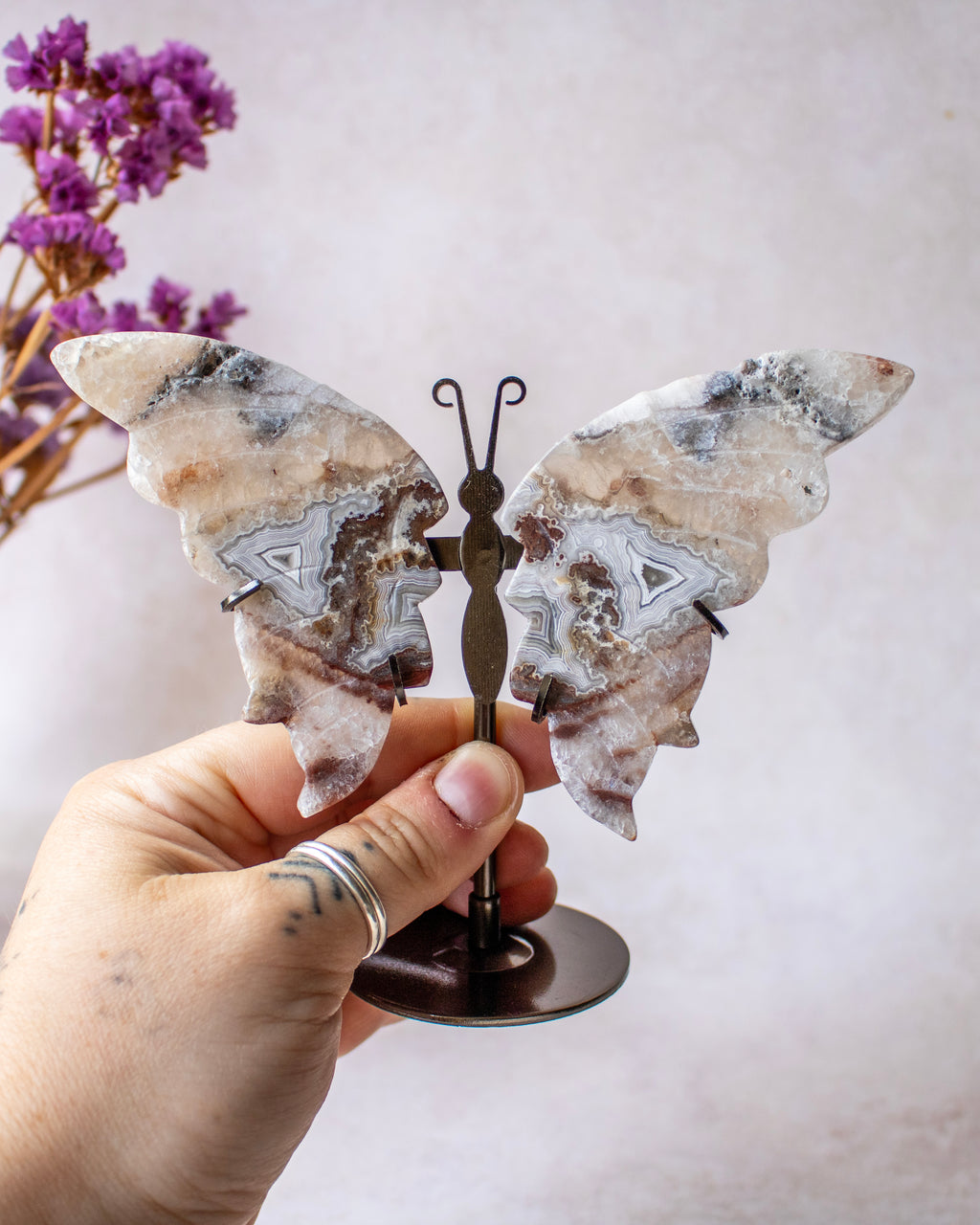 A hand holds a butterfly-shaped wall art piece made from polished agate stone wings with a small metal stand. The background is a soft beige wall with dried purple flowers in the upper left corner.