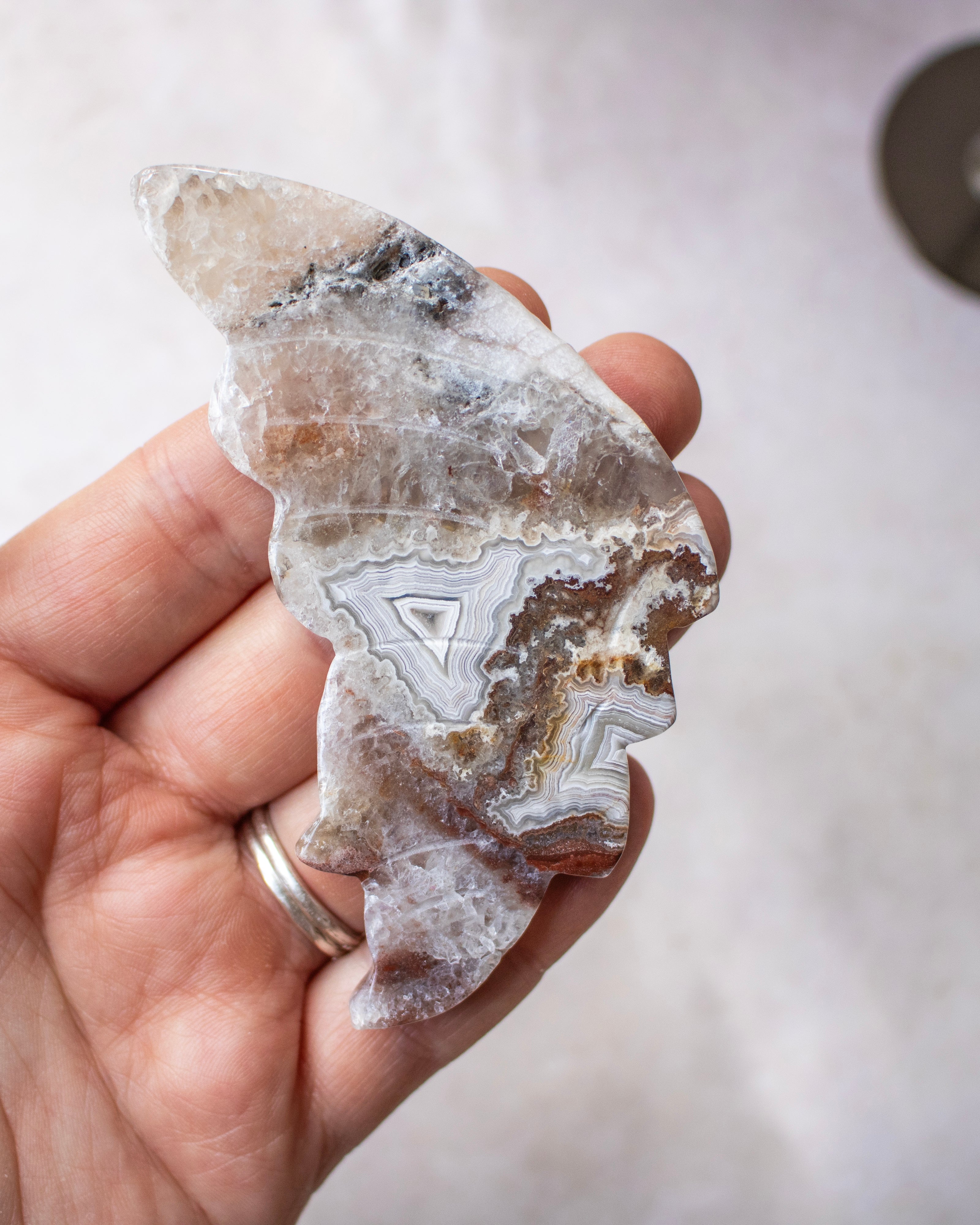 A hand holding a polished agate crystal cut in the shape of a wing silhouette, showcasing natural banded agate patterns in shades of gray, white, brown, and black.