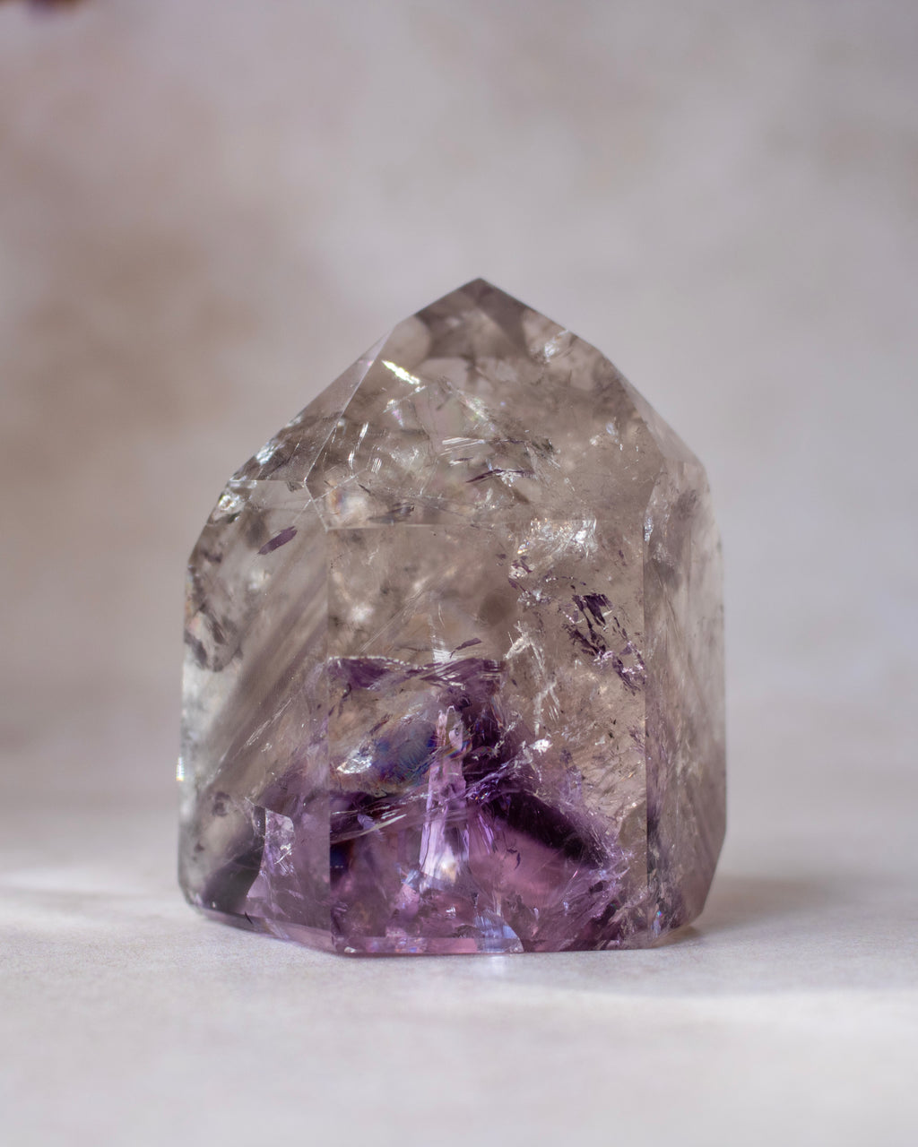 A natural clear quartz crystal point with prominent purple amethyst inclusions near the base, photographed on a soft neutral gray background.