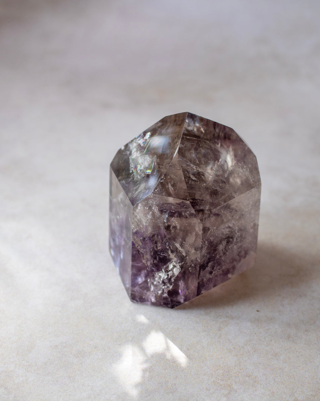 A faceted purple crystal gemstone, resembling amethyst, resting on a light beige textured surface.