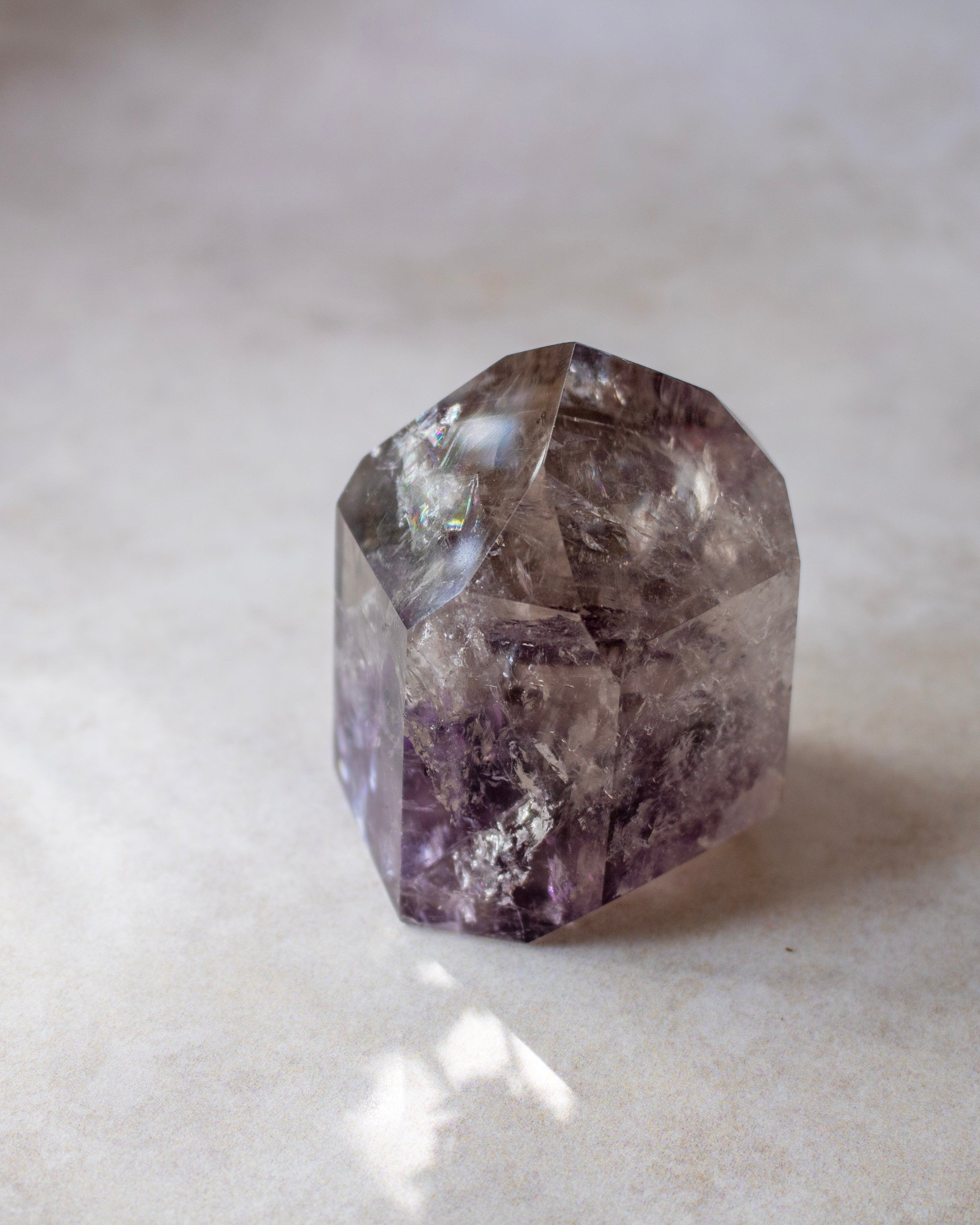A faceted purple crystal gemstone, resembling amethyst, resting on a light beige textured surface.