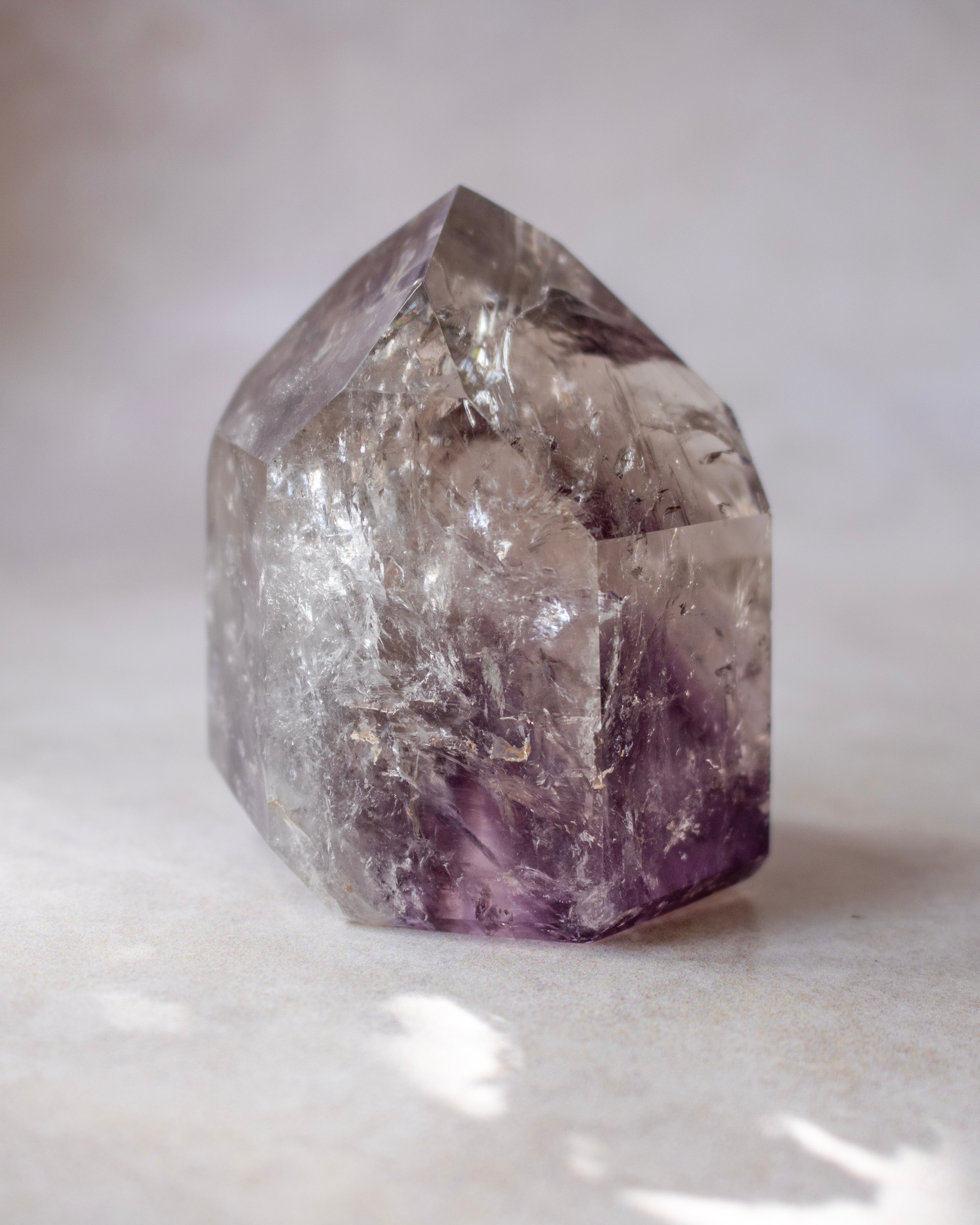 A natural amethyst crystal point with a hexagonal shape, showing clear faceted transparency with deep purple hues concentrated towards the base, set against a soft, neutral gray background.