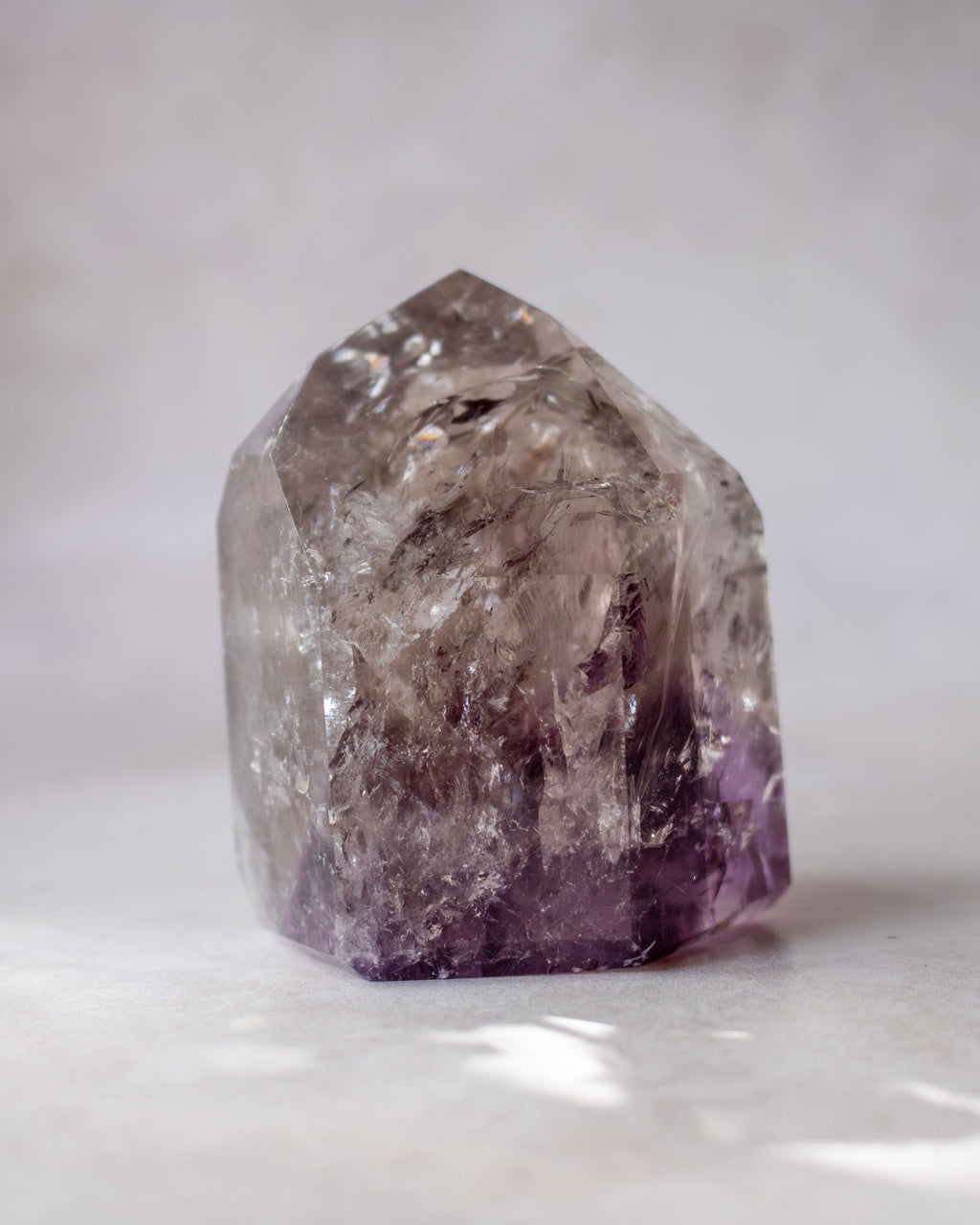 A raw amethyst crystal cluster with natural geometric facets, showing a gradient from translucent smoky gray at the top to deep purple at the base, photographed on a neutral light gray surface.