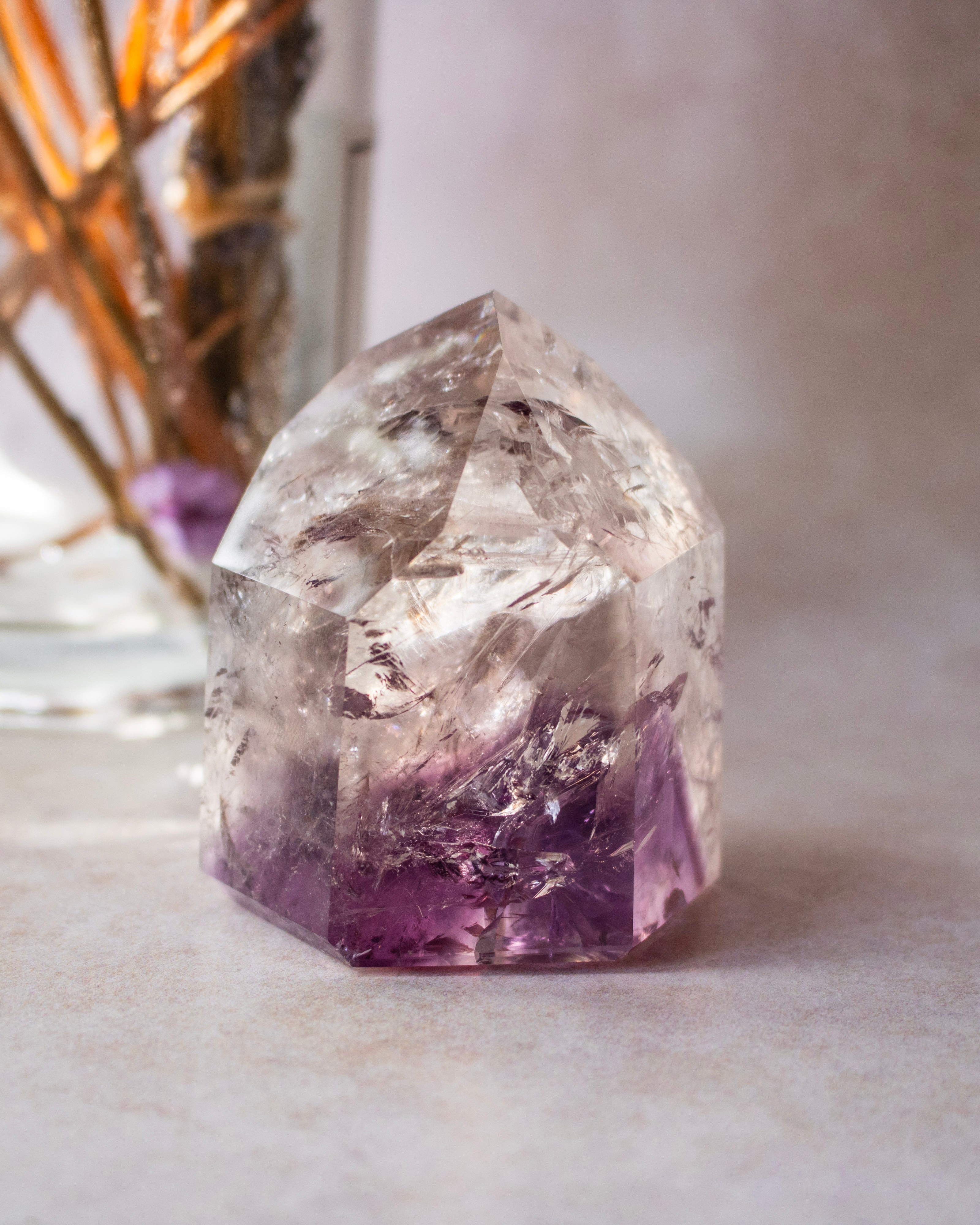 A natural clear quartz crystal point with a purple amethyst base, displayed on a neutral surface with a blurred decorative background.
