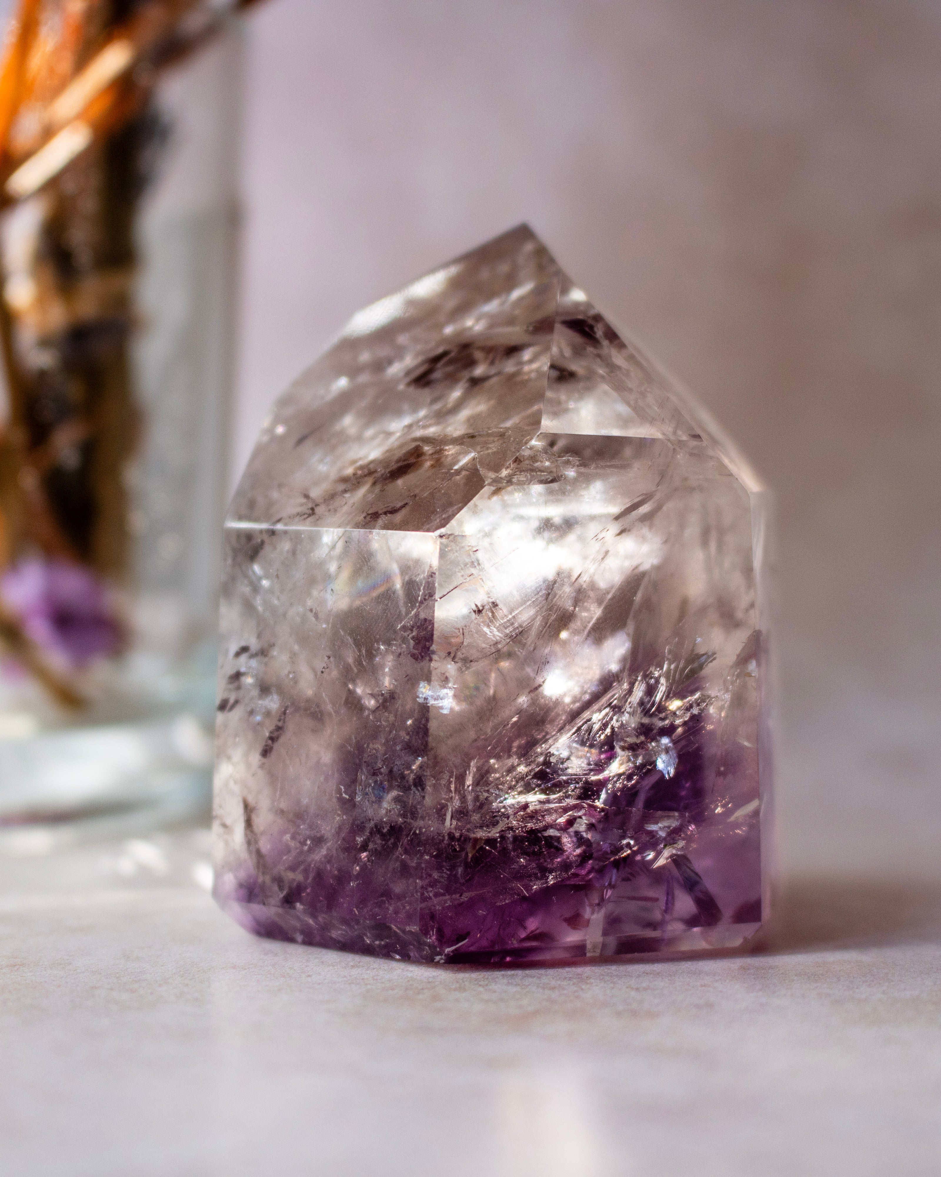 A natural amethyst crystal tower with a pointed top, showing a gradient from clear and smoky gray at the top to deep purple at the base, set against a softly blurred neutral background.