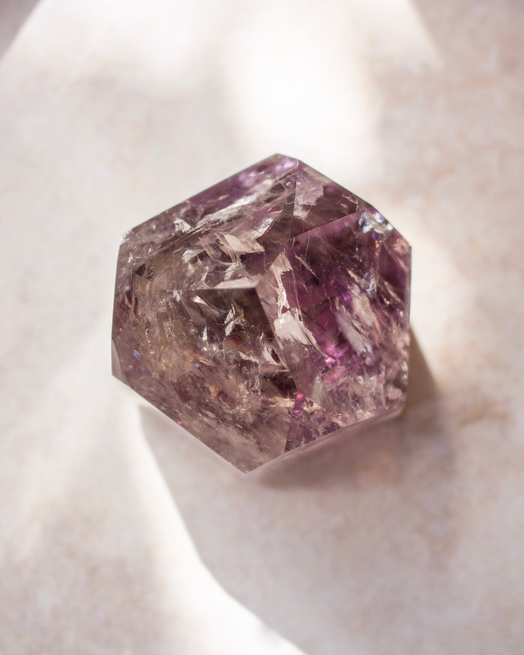 A faceted purple crystal gemstone with natural inclusions, resting on a textured beige surface with soft shadows.