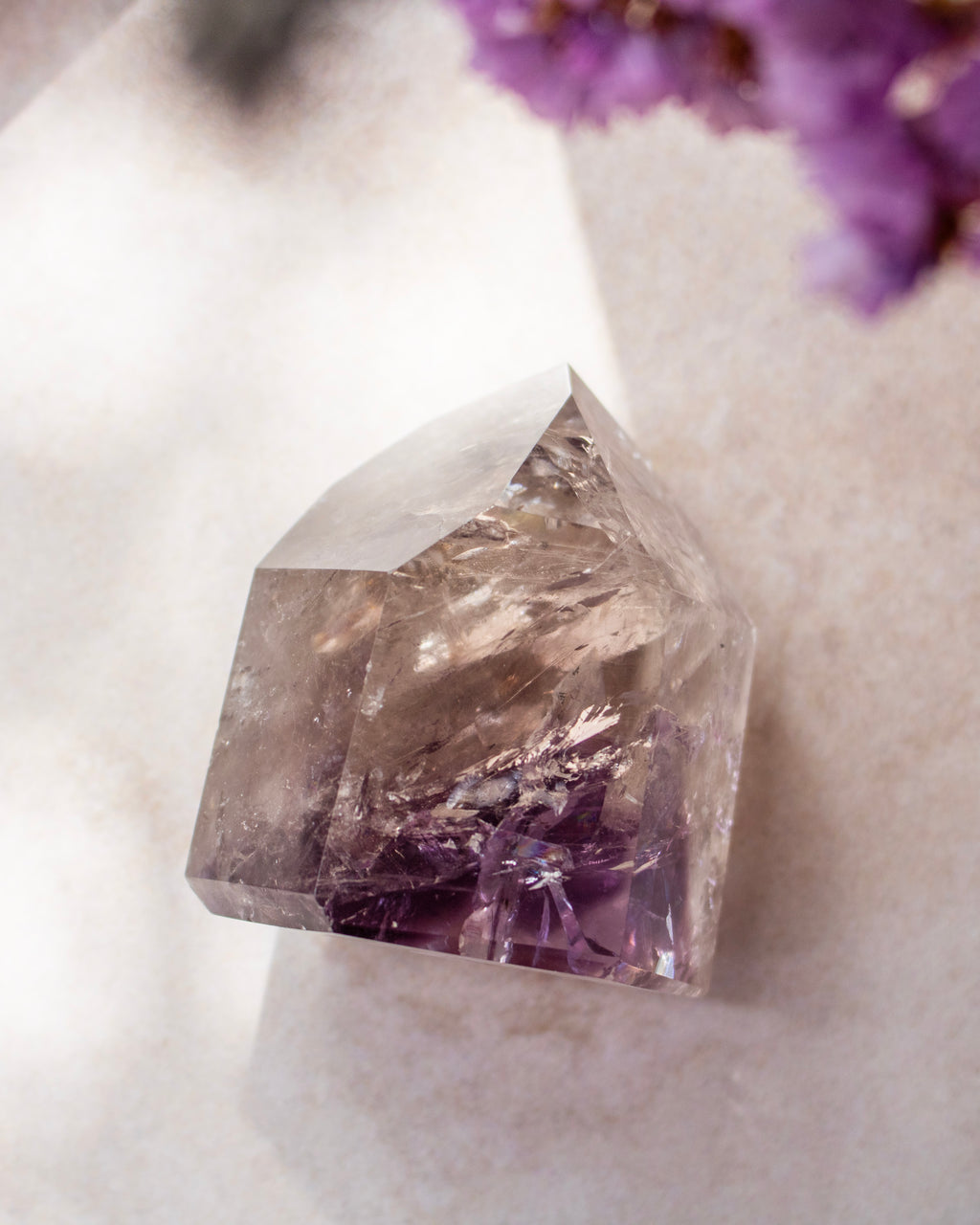A natural pointed crystal cluster, likely amethyst, showing clear faceted transparency with deep purple inclusions near the base, photographed on a soft light surface with blurred purple flowers in the corner.