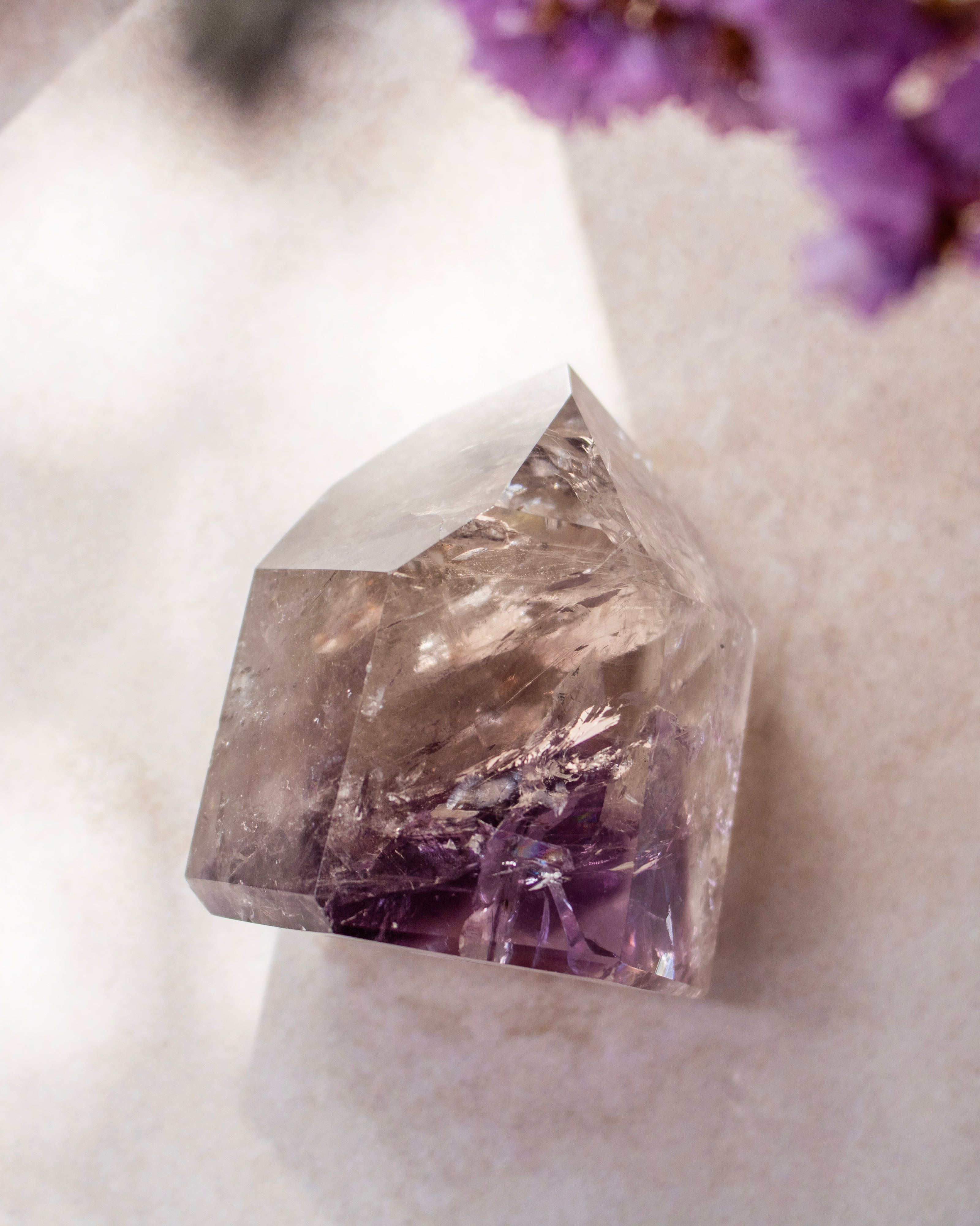 A natural pointed crystal cluster, likely amethyst, showing clear faceted transparency with deep purple inclusions near the base, photographed on a soft light surface with blurred purple flowers in the corner.