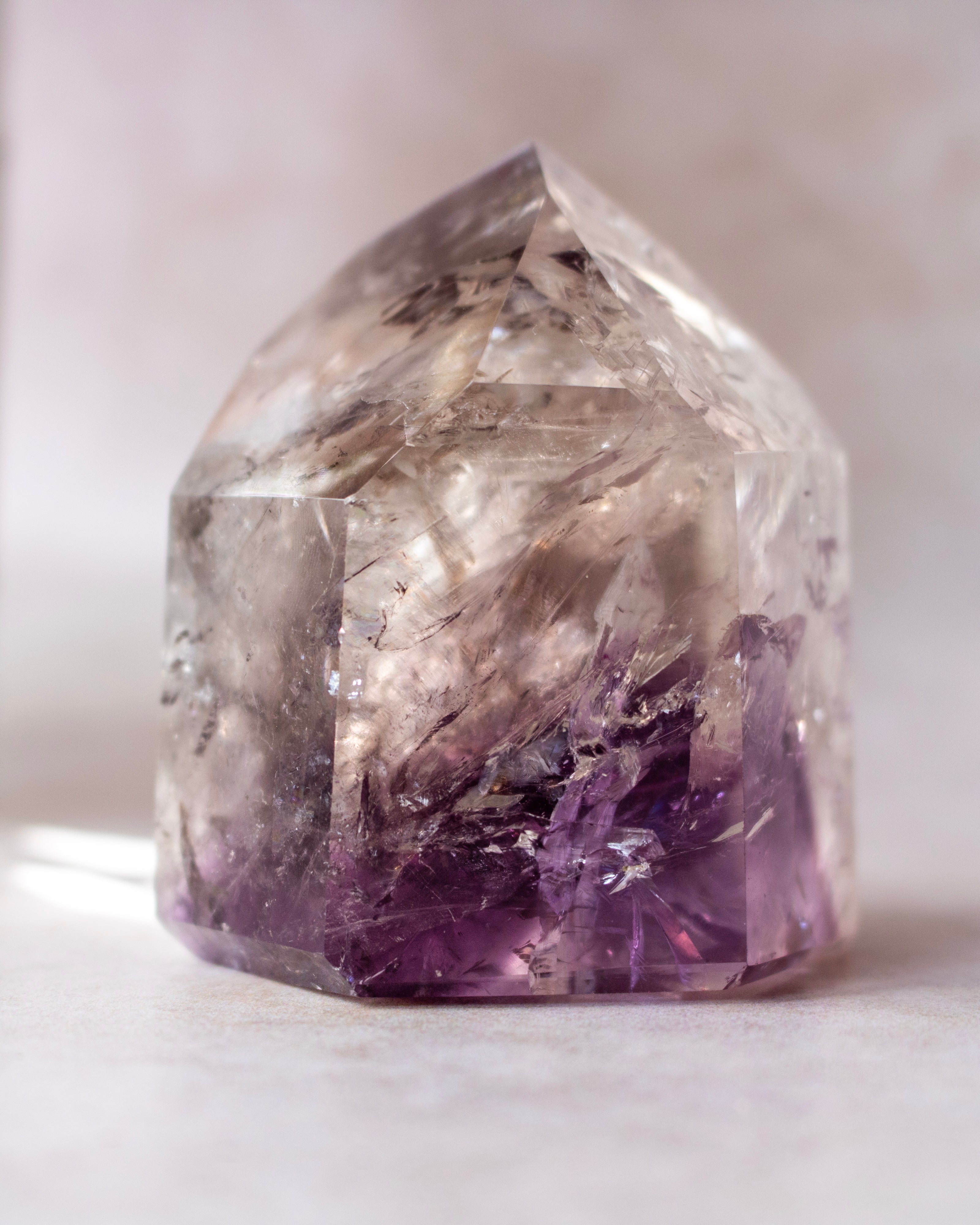 A clear quartz crystal point with prominent purple amethyst inclusions concentrated near the base, photographed on a soft neutral background.
