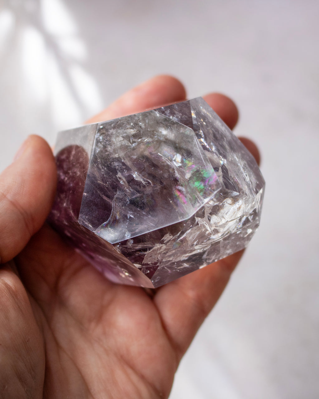 A hand holding a faceted clear crystal with subtle purple and iridescent hints, photographed against a softly blurred light background.