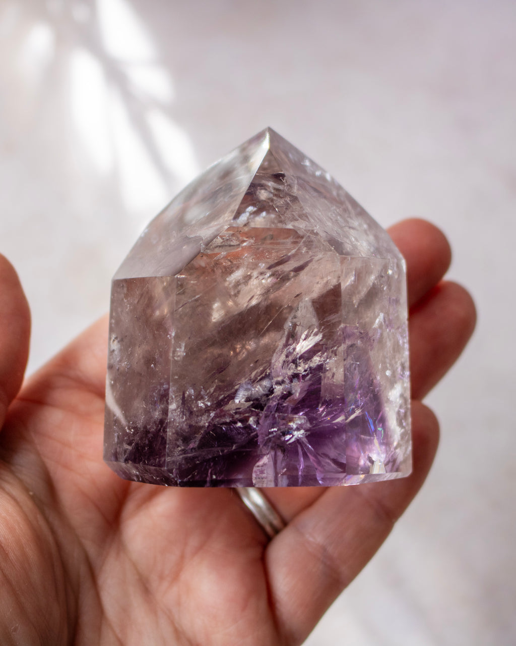A house-shaped clear quartz crystal with purple amethyst inclusions, held in a person's hand against a soft, light background.