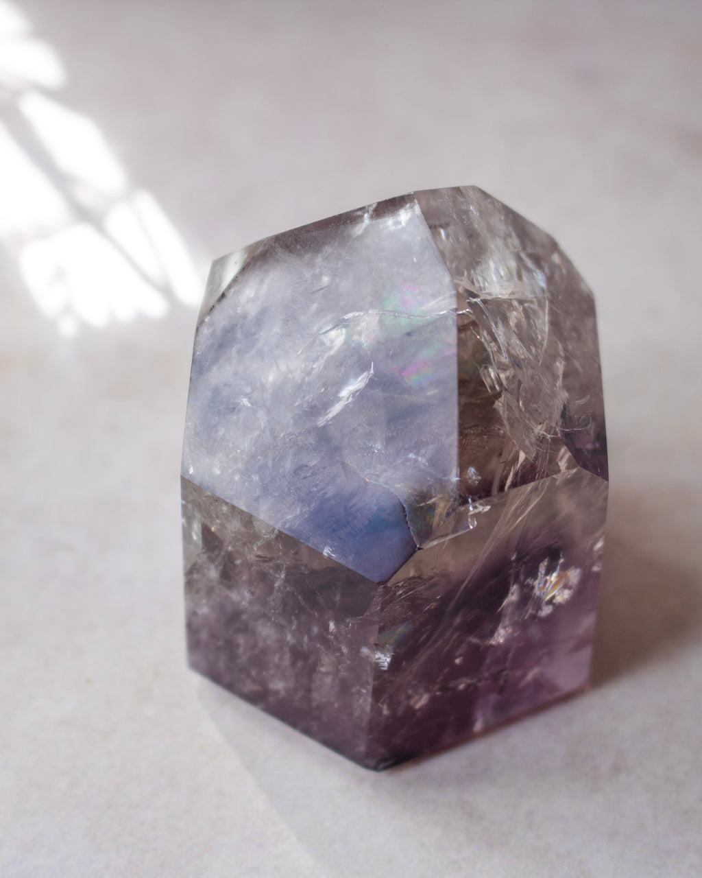 A faceted raw crystal stone with a hexagonal shape, showing natural mineral inclusions and color variations from light gray to deep purple, photographed on a neutral light gray surface.