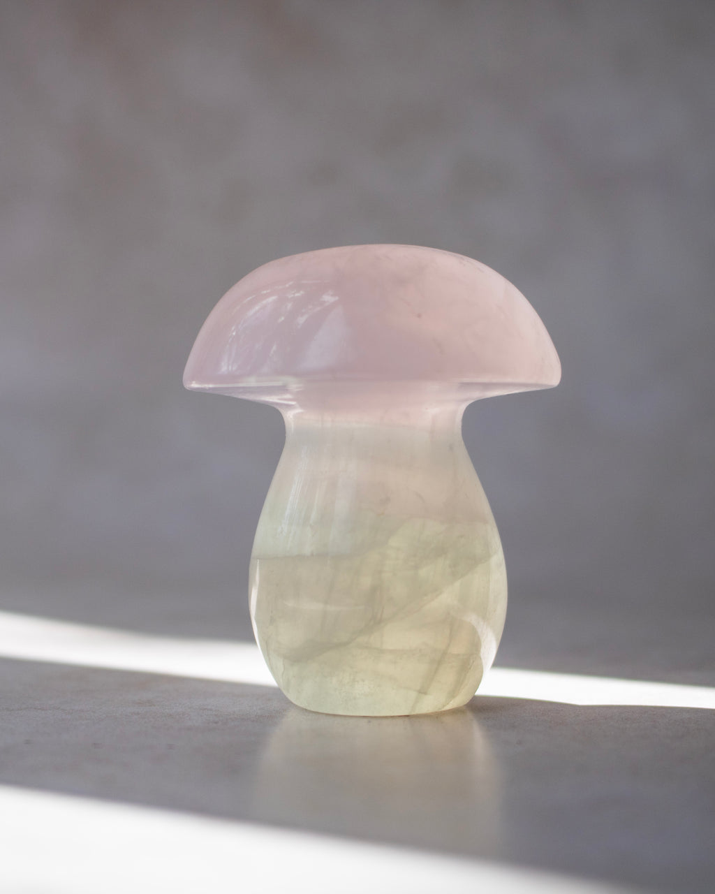 A small mushroom-shaped decorative object carved from pale pink and milky translucent stone, standing on a gray surface against a softly blurred gray background.