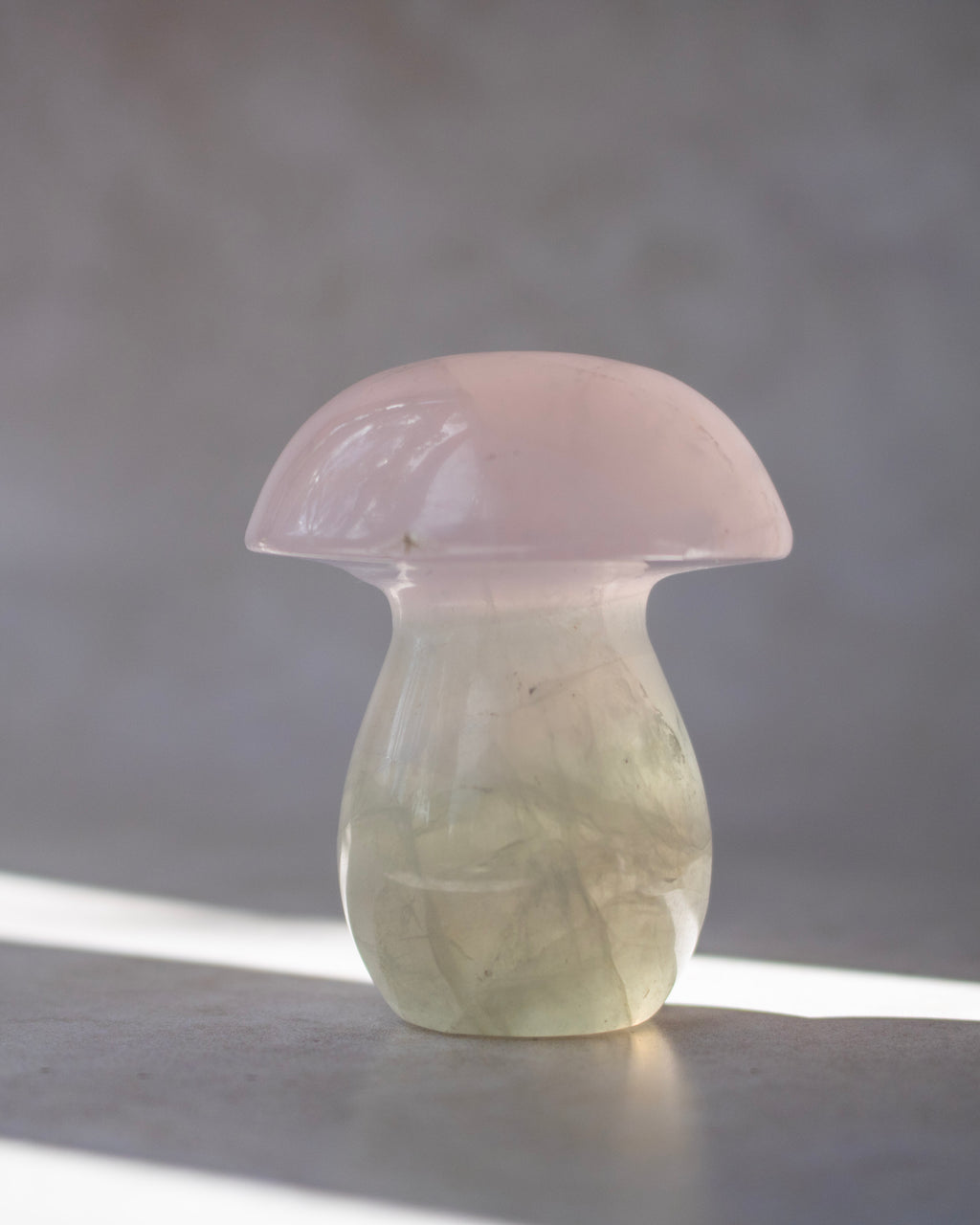 A small mushroom-shaped decorative object carved from pale pink and milky white stone, standing on a gray surface with soft natural light and a blurred gray background.