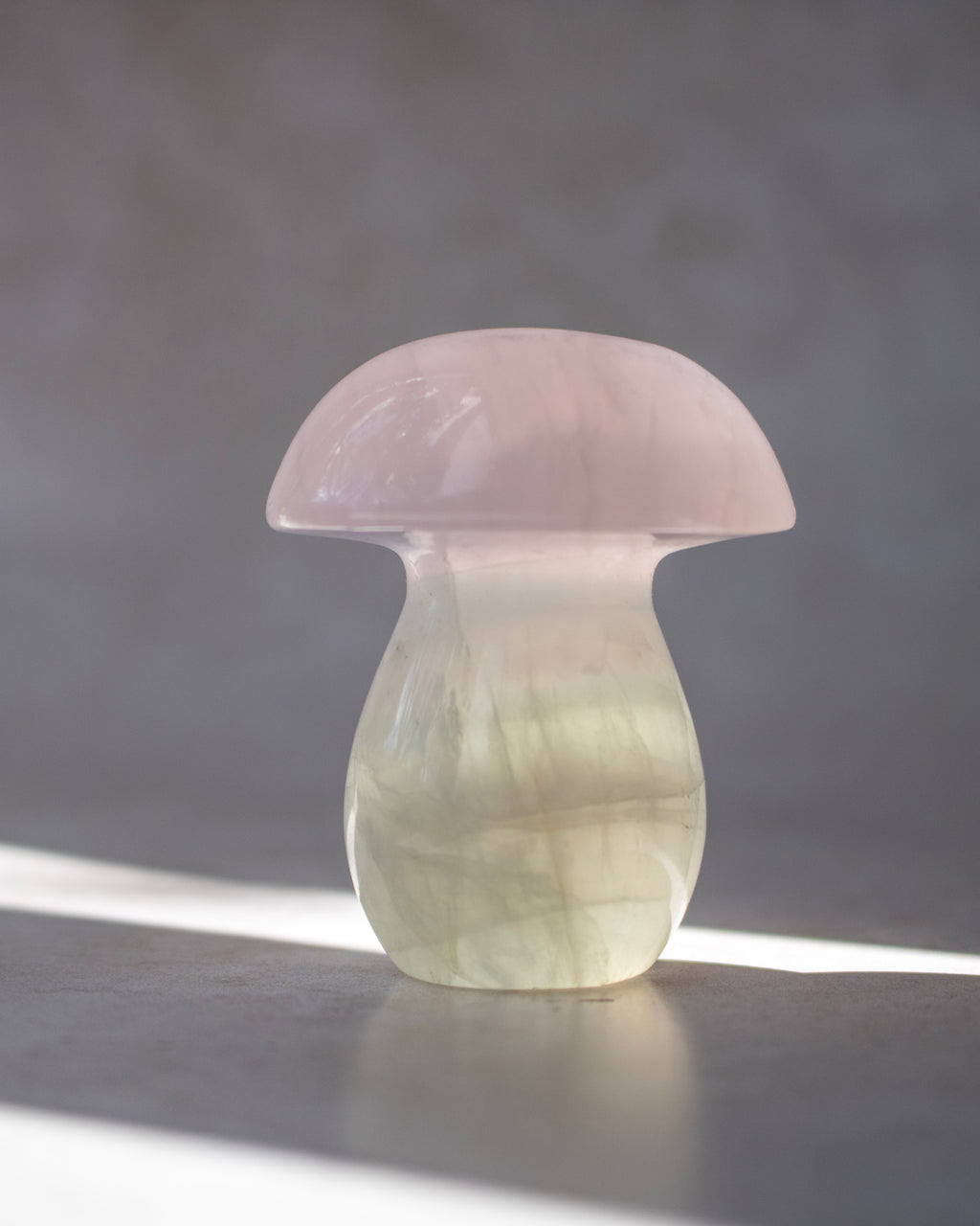 A mushroom-shaped decorative object made of translucent pale pink and milky white stone, standing on a gray surface against a gray background with a bright light reflection.