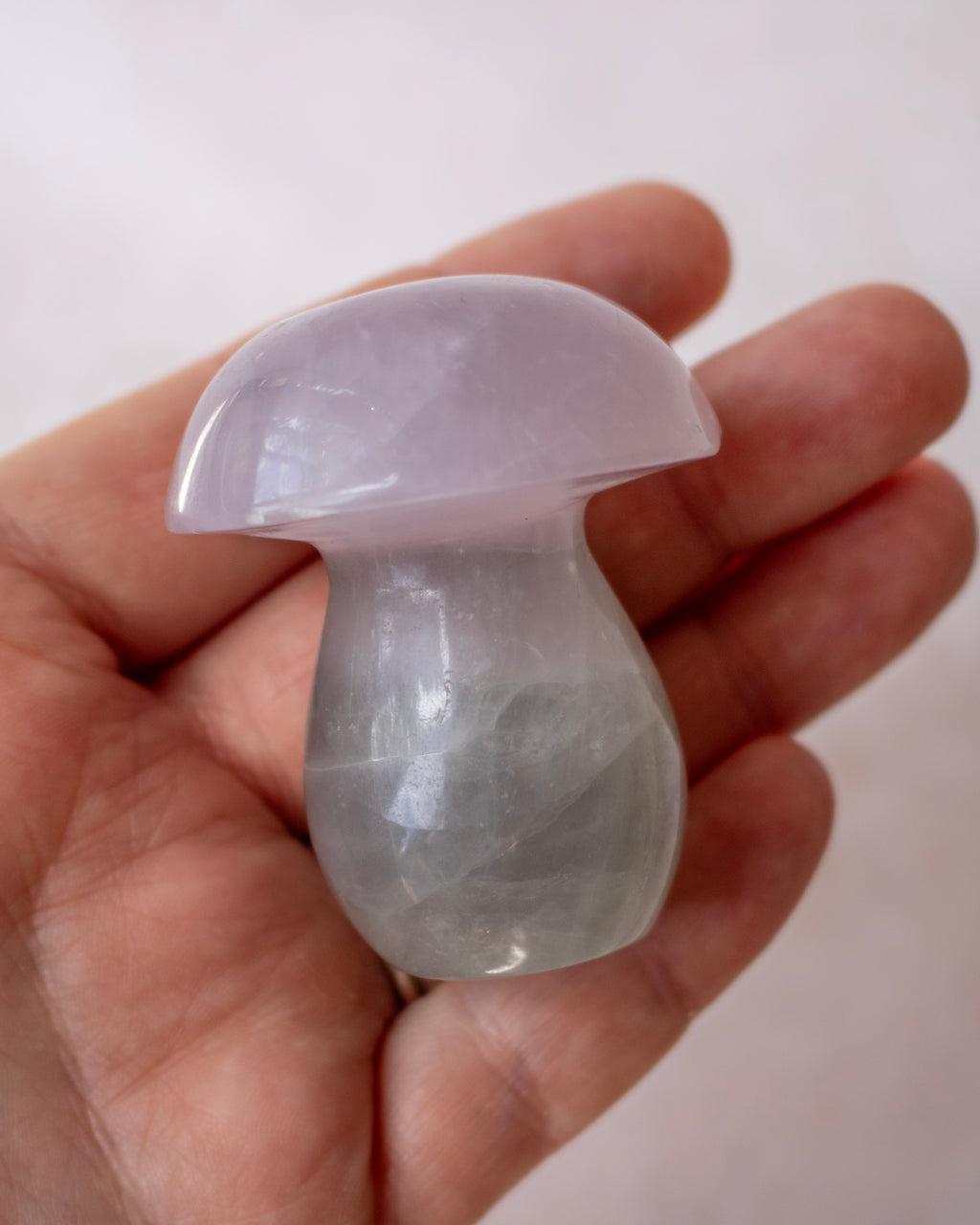 A small polished mushroom-shaped crystal gemstone in pale lavender and gray tones, held in a person's hand against a soft light gray background.