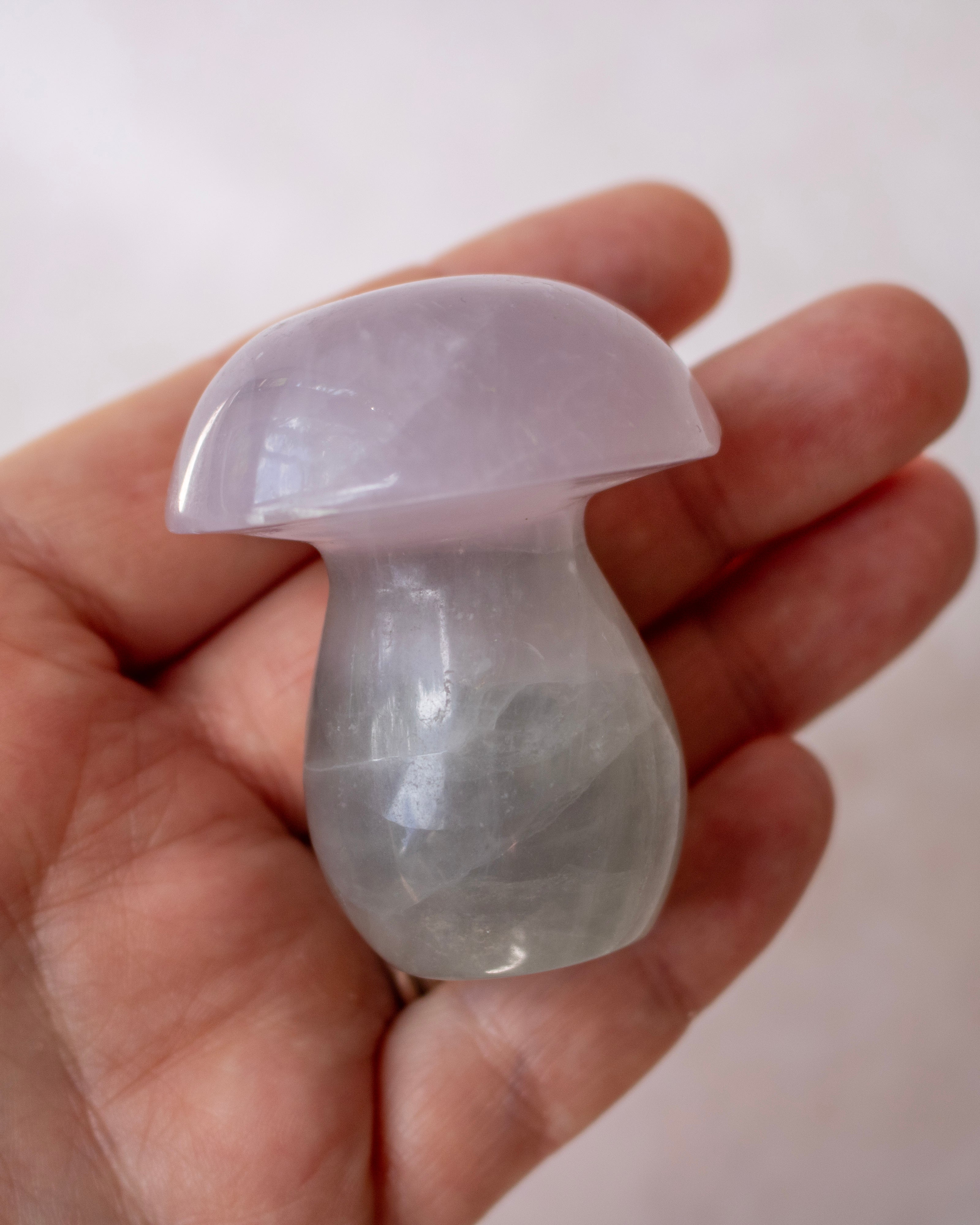 A small polished mushroom-shaped crystal gemstone in pale lavender and gray tones, held in a person's hand against a soft light gray background.