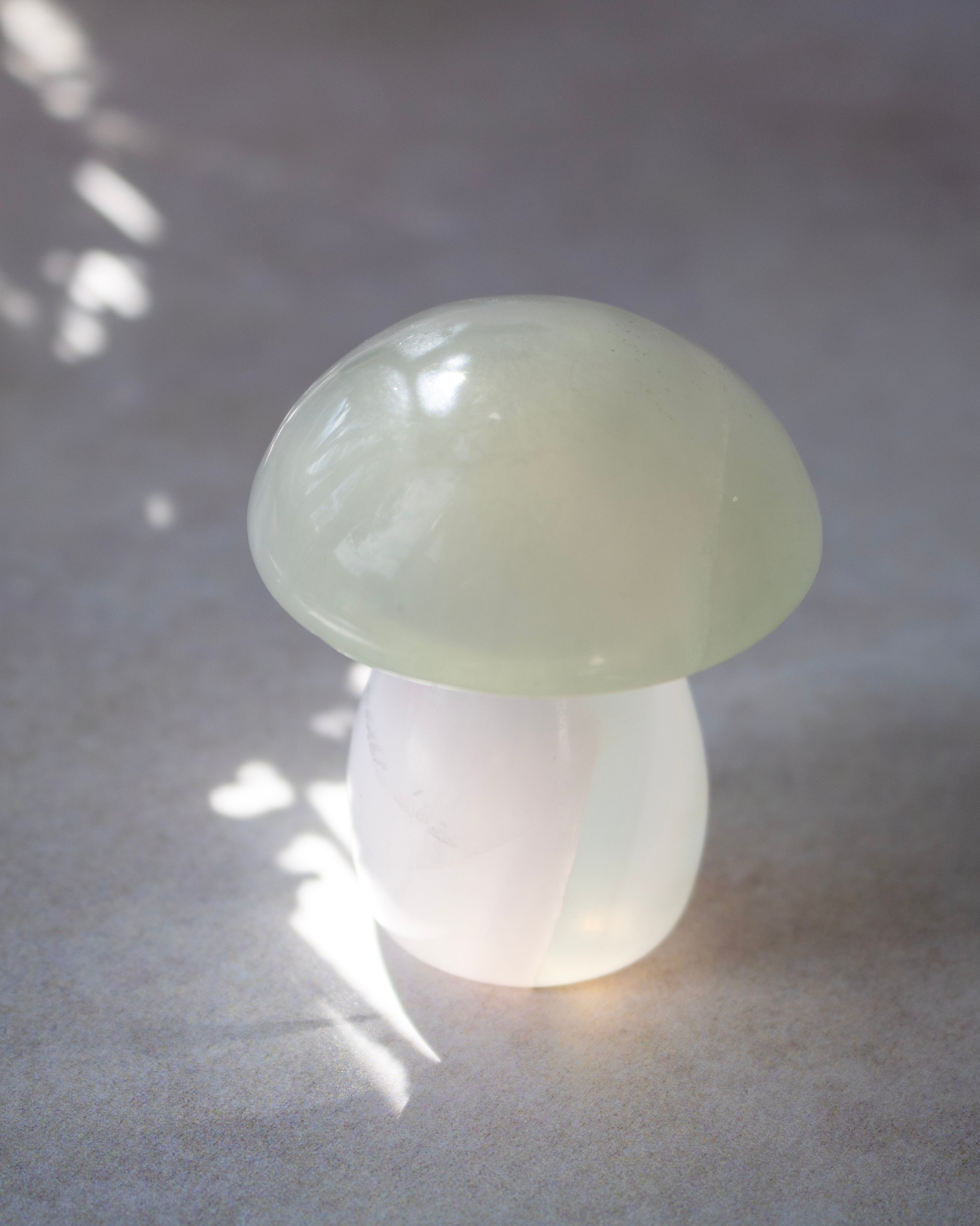 A small mushroom-shaped decorative object made of smooth translucent pale green and white stone, photographed on a gray surface with soft leaf shadows.