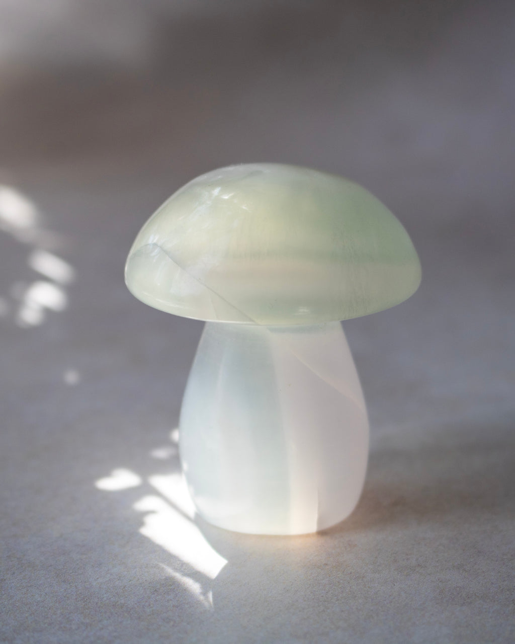 A small mushroom-shaped decorative object made of milky translucent material, photographed on a soft gray surface with dappled light and shadow.
