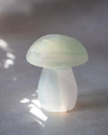 A small mushroom-shaped decorative object made of milky translucent material, photographed on a soft gray surface with dappled light and shadow.