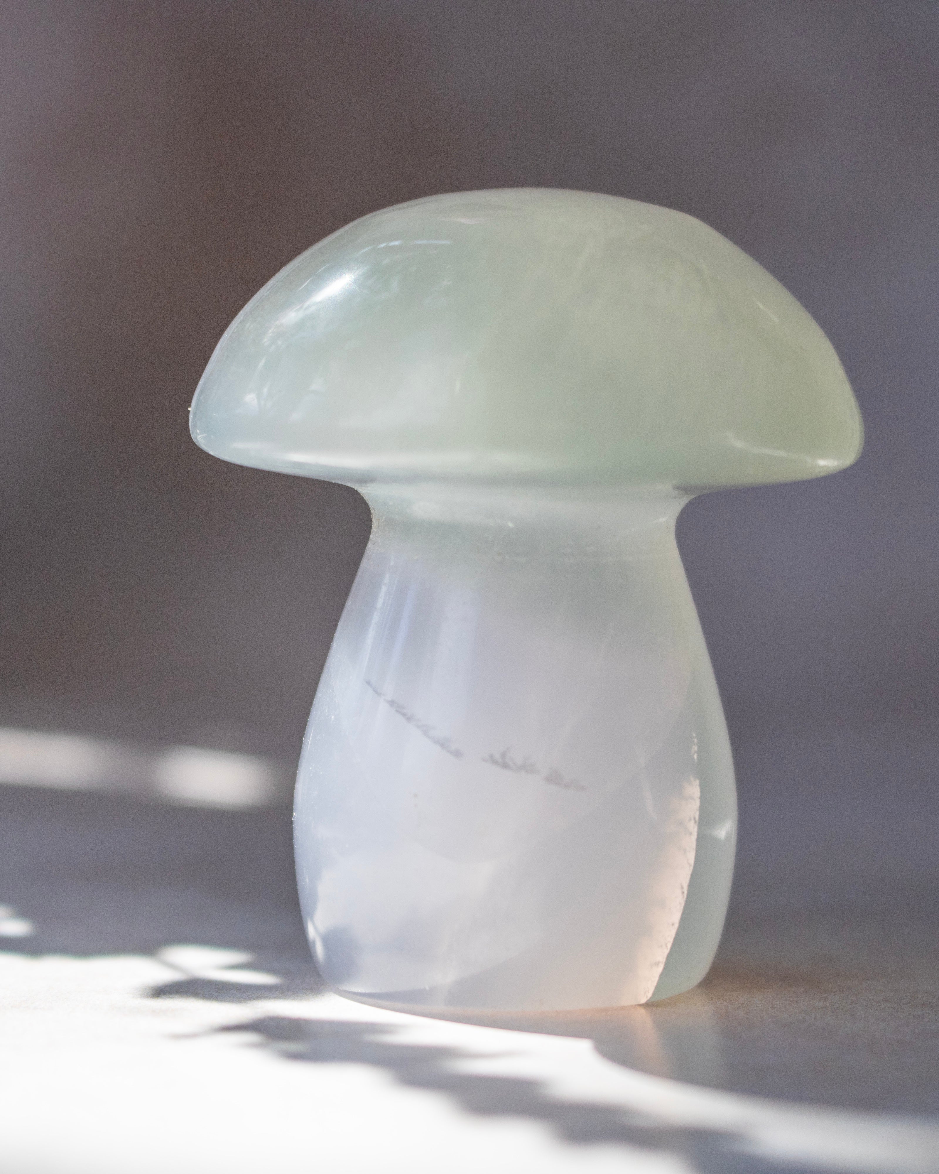A mushroom-shaped translucent stone sculpture, made from pale greenish-white agate, standing on a light surface with soft shadows and bright light filtering from the side.