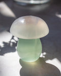 A small mushroom-shaped decorative object made of smooth translucent pale green and milky white material, photographed on a gray surface with dappled sunlight and shadows.