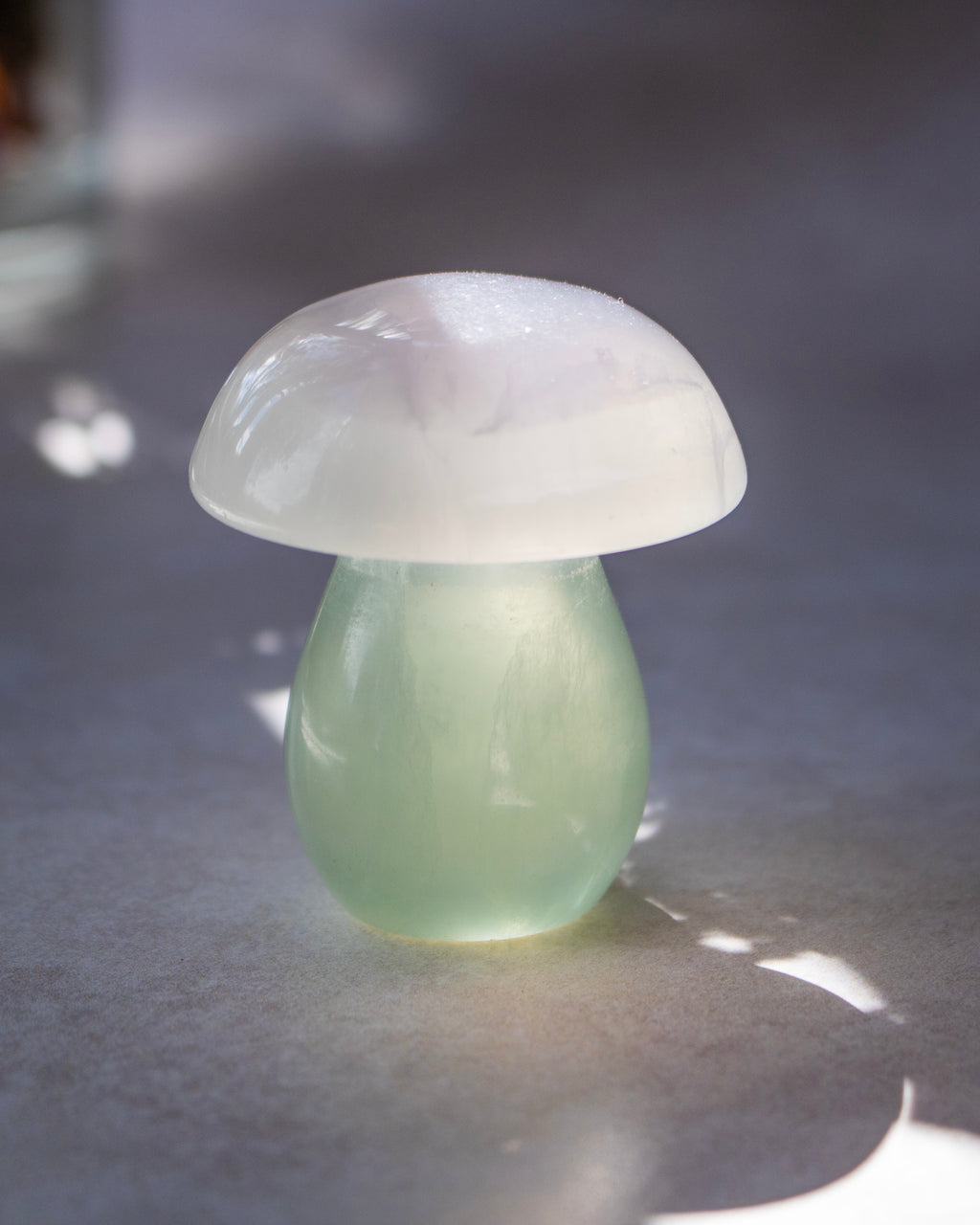 A small mushroom-shaped decorative object made of smooth translucent stone, with a pale green stem and a milky white cap, photographed on a gray surface with soft natural shadows.