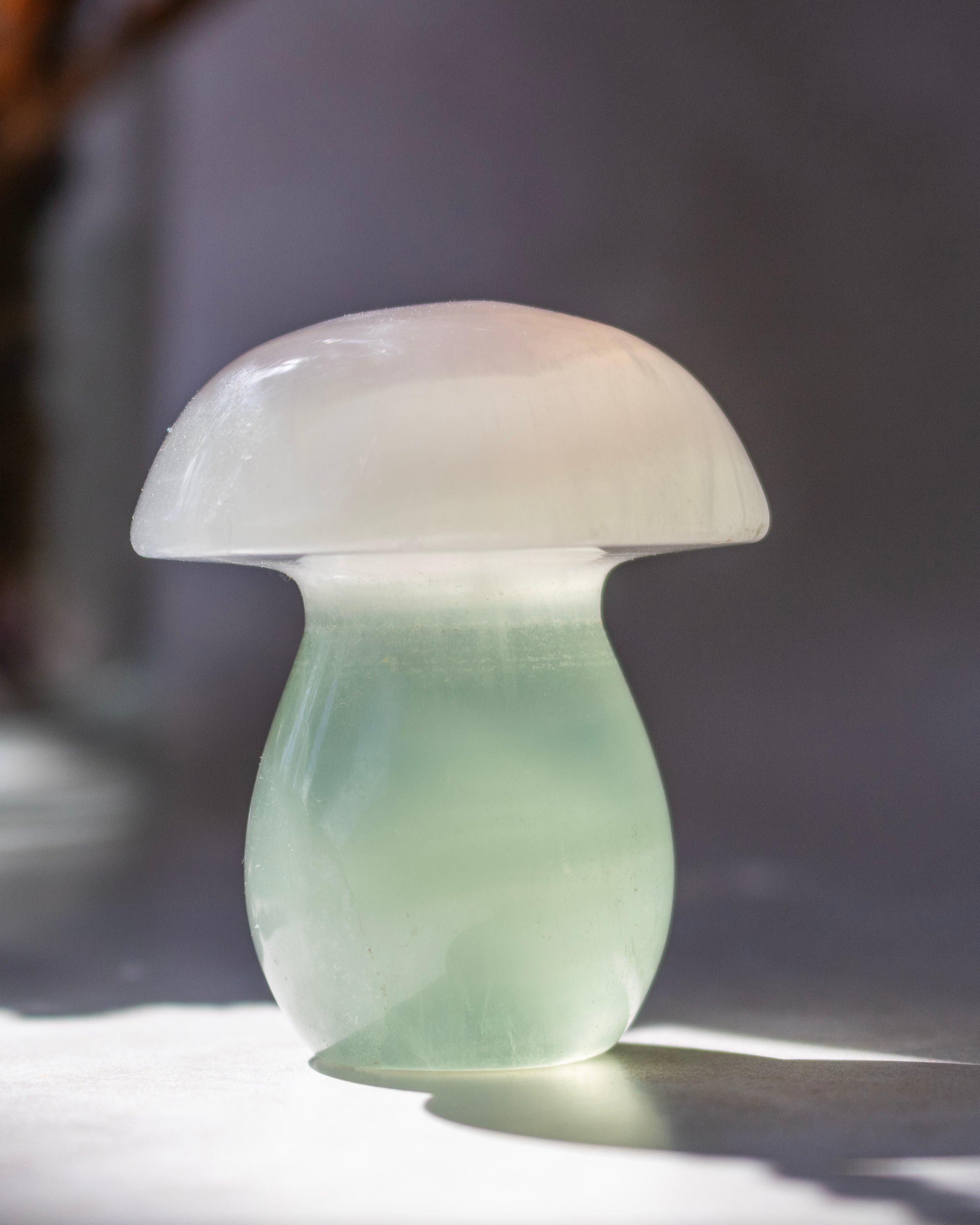 A mushroom-shaped translucent decorative object made of pale green and milky white material, photographed on a light surface with soft natural light and a blurred gray background.