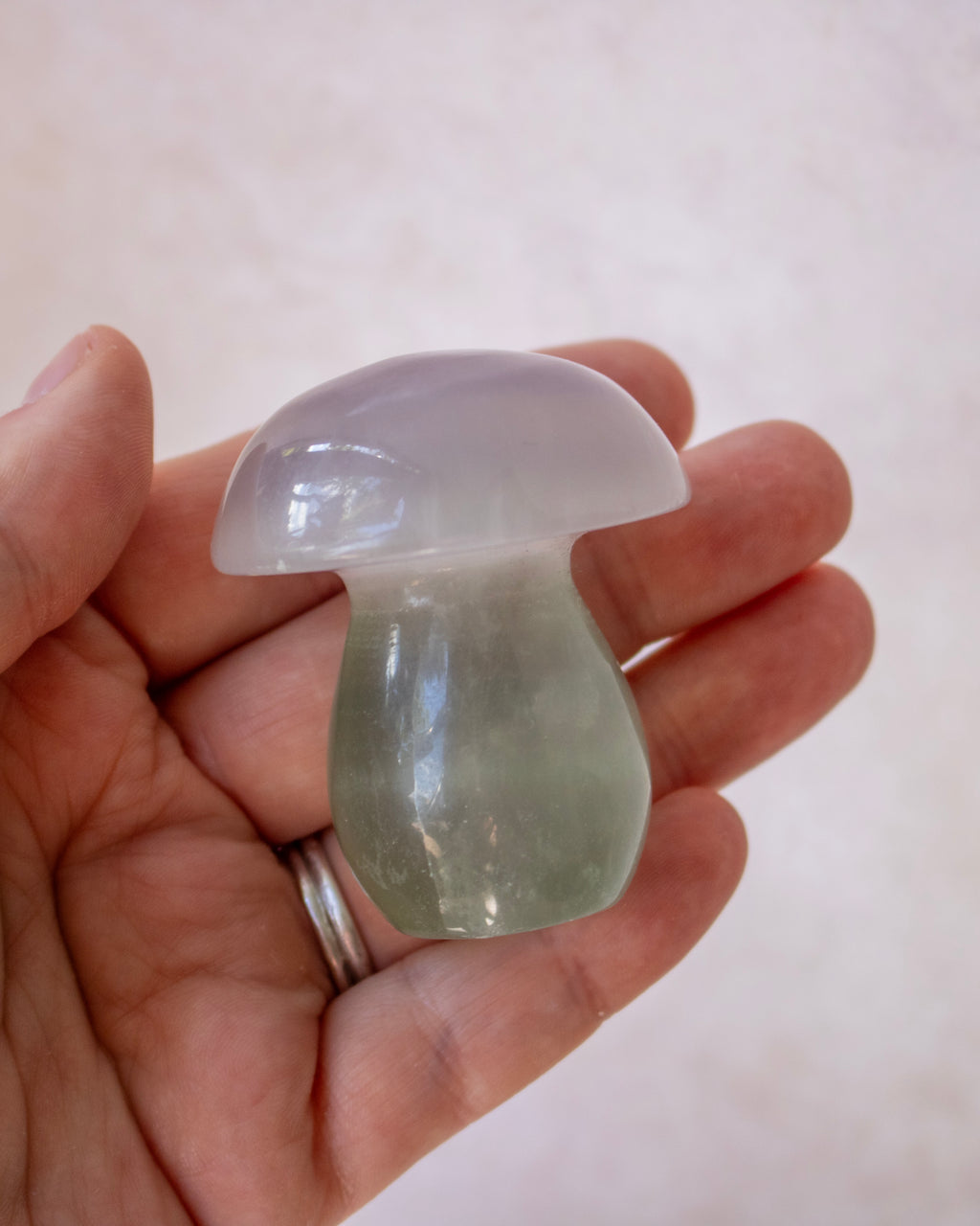 A hand holding a small mushroom-shaped translucent stone ornament, showing a gradient from pale lavender-gray on top to light green at the base, against a soft beige background.