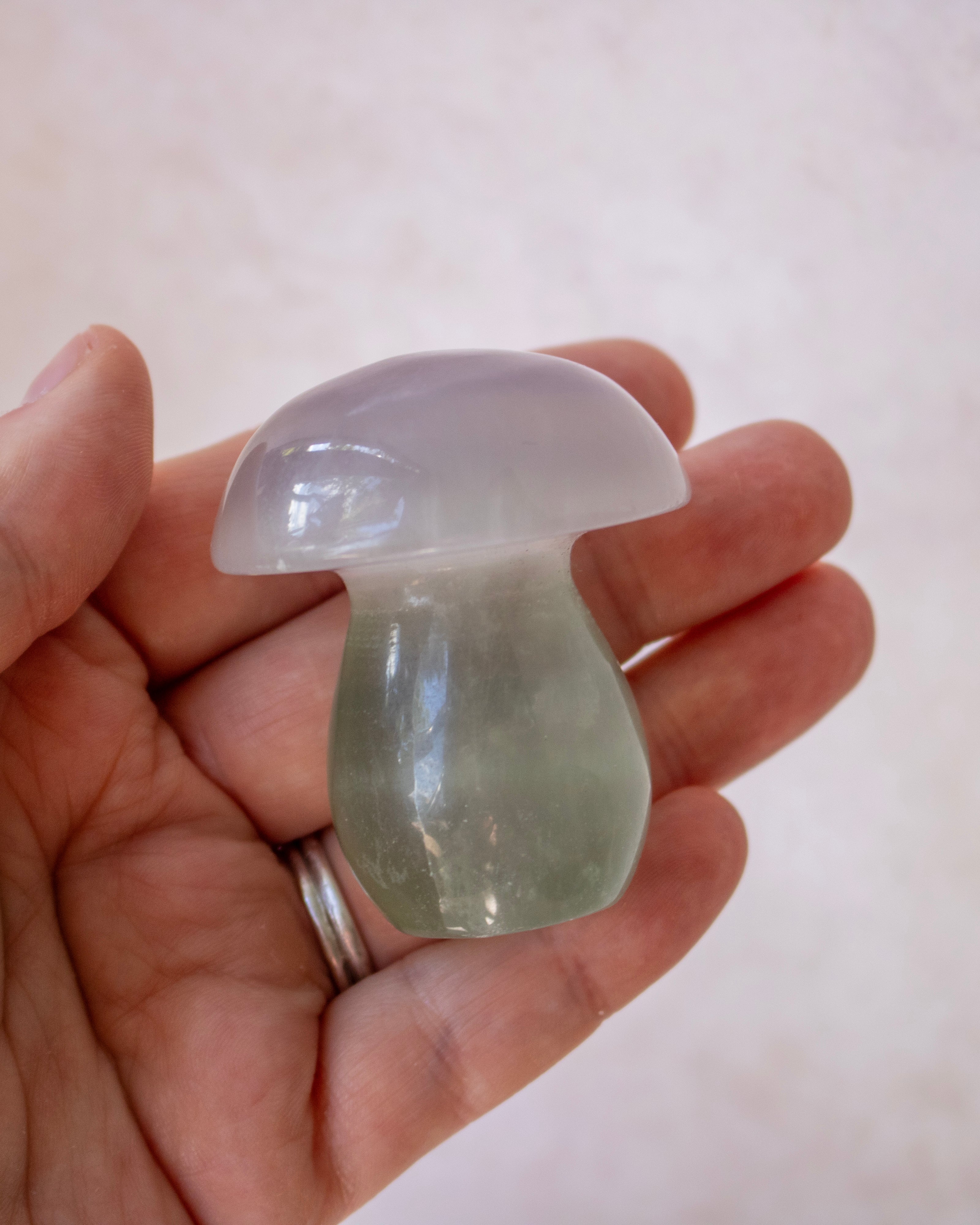 A hand holding a small mushroom-shaped translucent stone ornament, showing a gradient from pale lavender-gray on top to light green at the base, against a soft beige background.